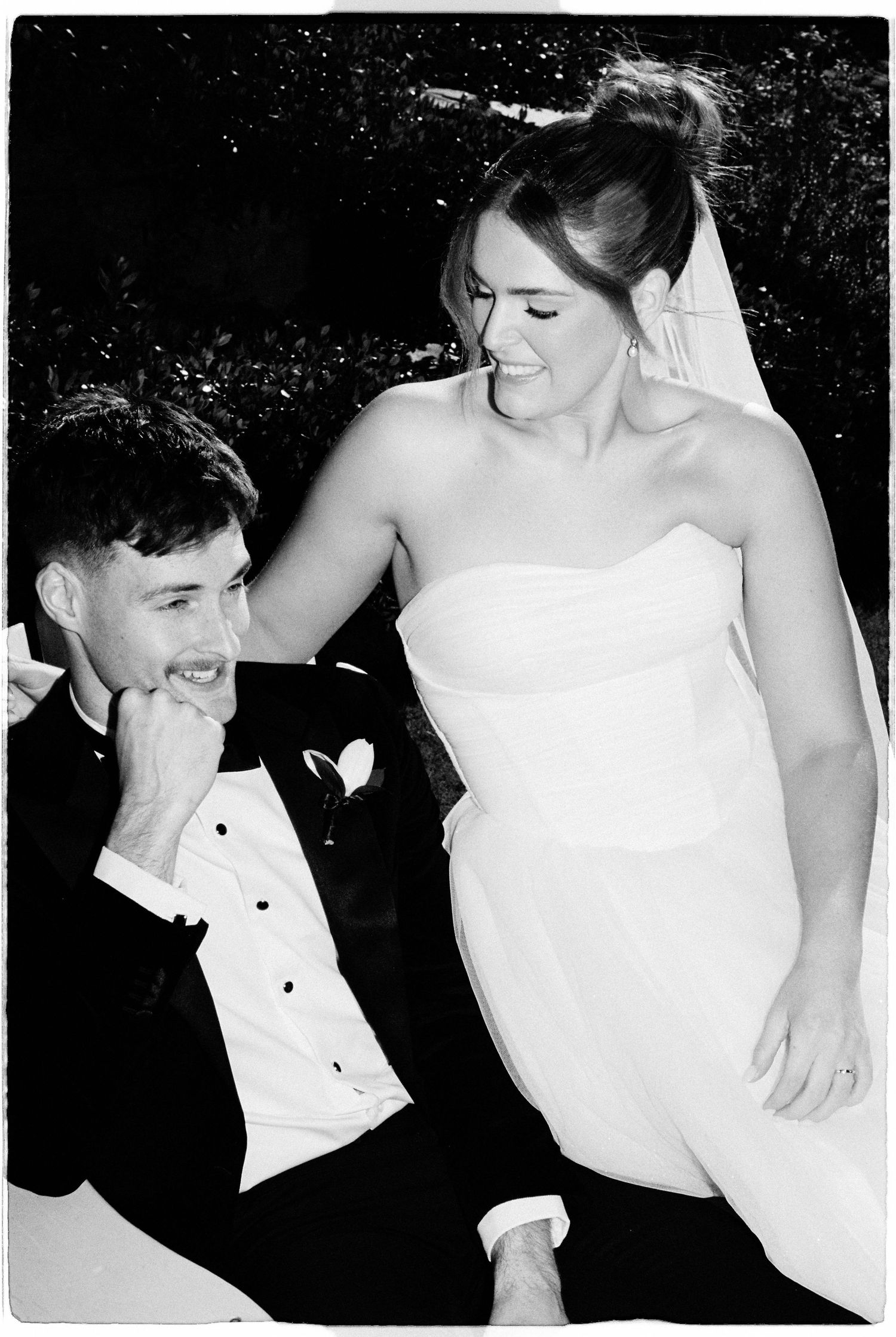 Black and white wedding photo of a smiling bride in strapless gown and groom in tuxedo sharing a joyful moment.