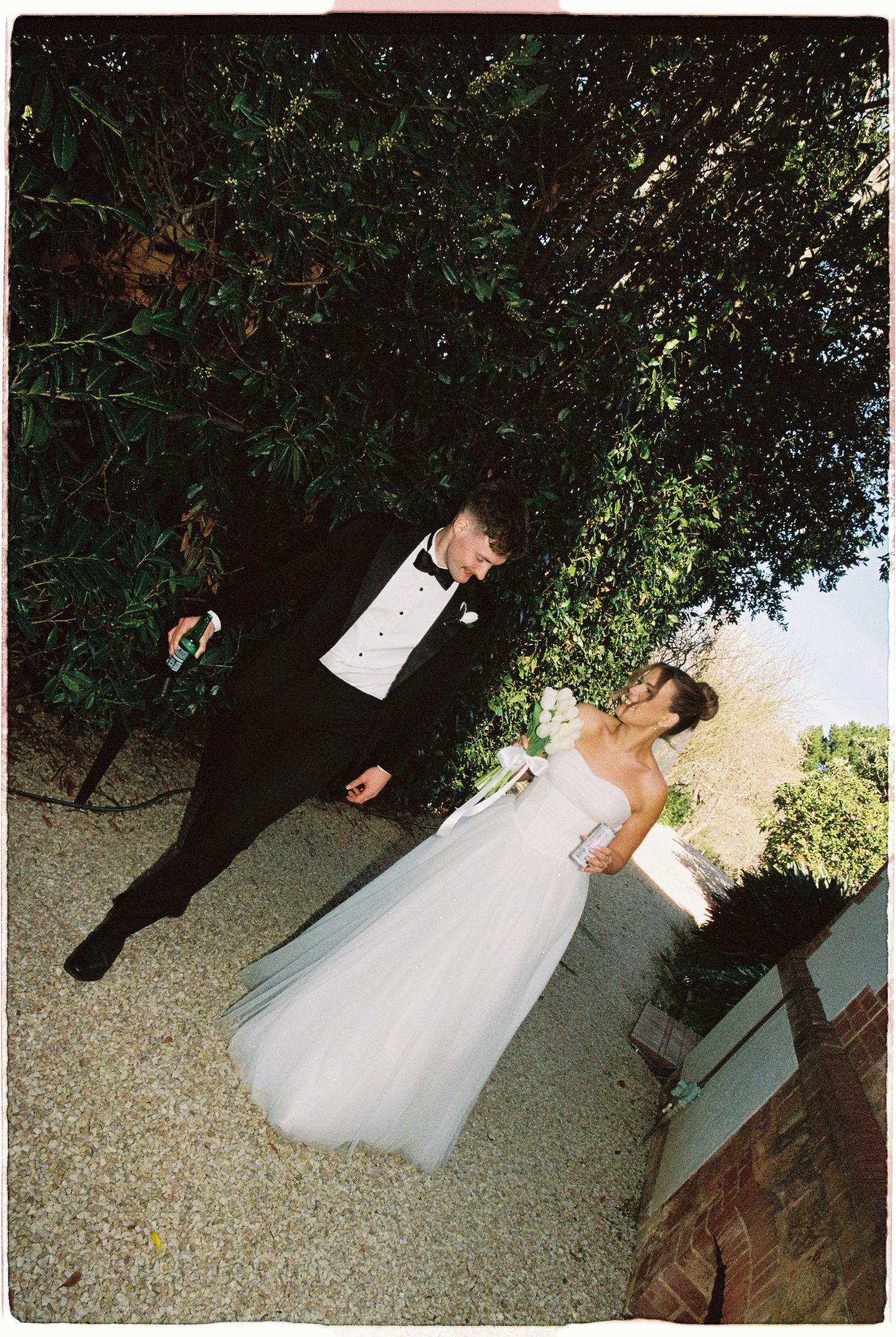 A bride in a white gown and groom in a black tuxedo share a romantic moment outdoors near lush green hedges.