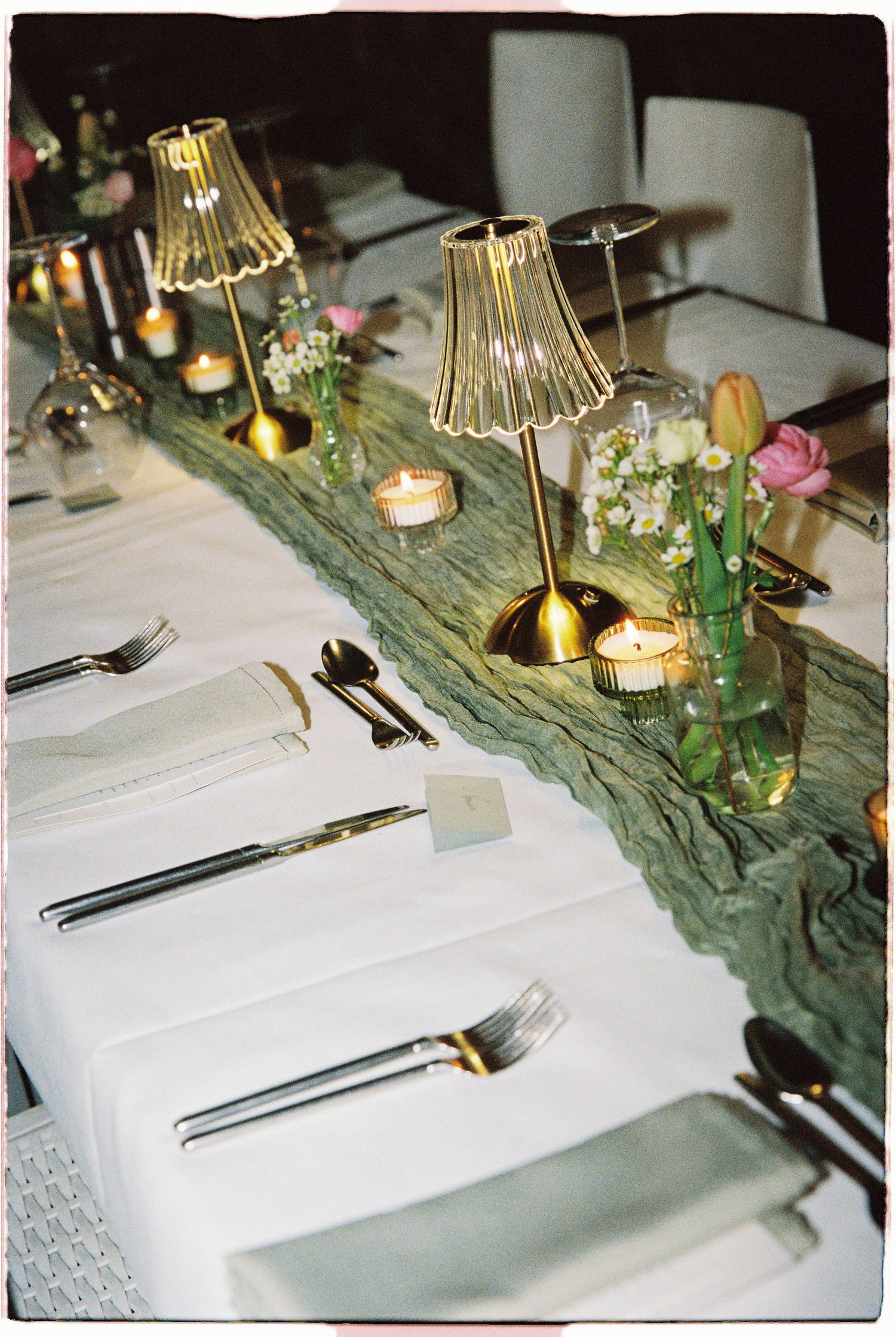 An elegantly set wedding reception table with gold lamp centerpieces, candles, fresh flowers, and white linens.