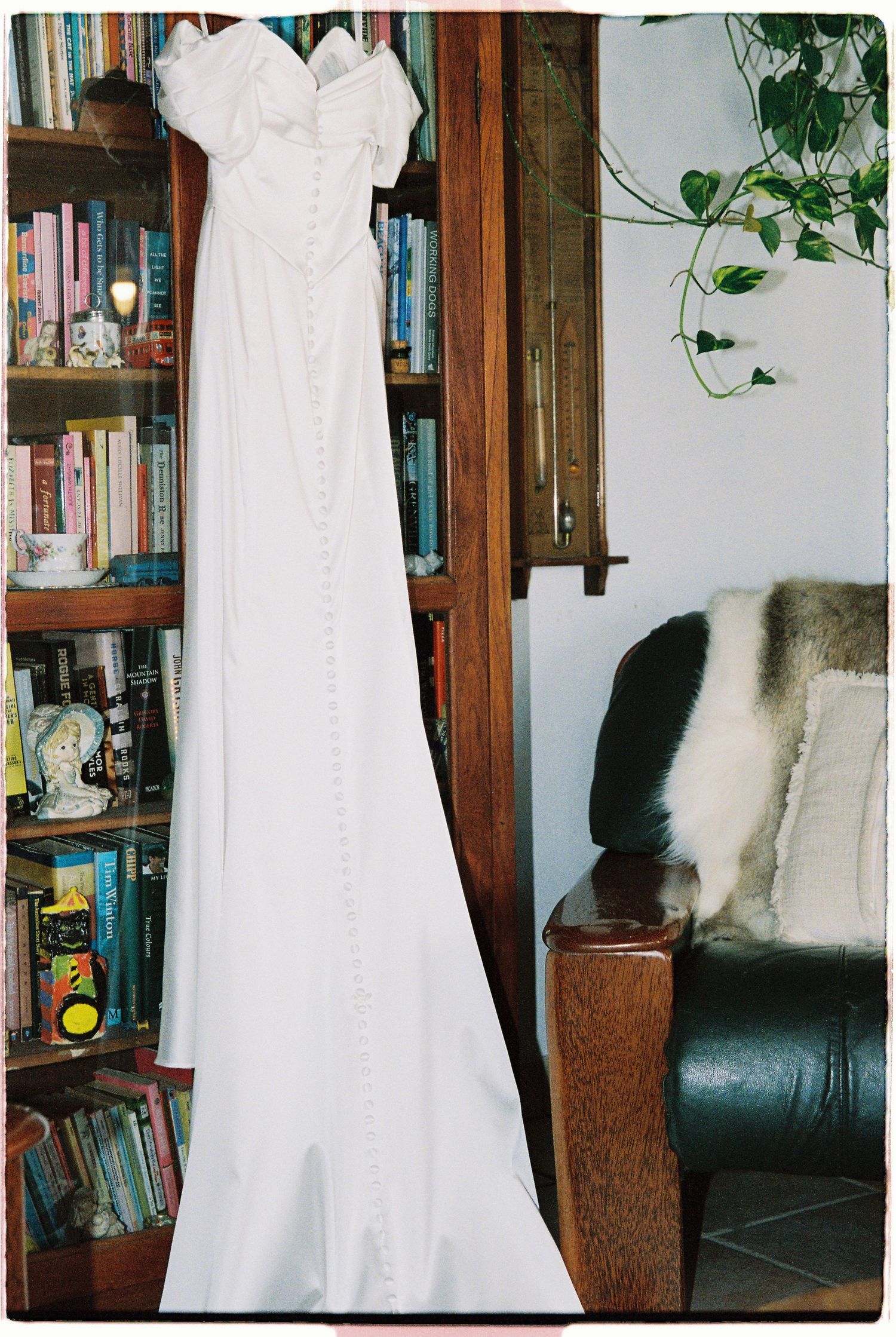 Elegant white bridal gown with button detail hanging beside bookshelf and leather chair.