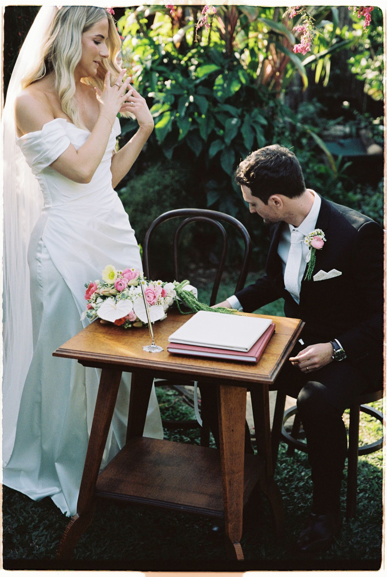 Bride and groom signing wedding register at wooden table with floral bouquet in lush garden setting.
