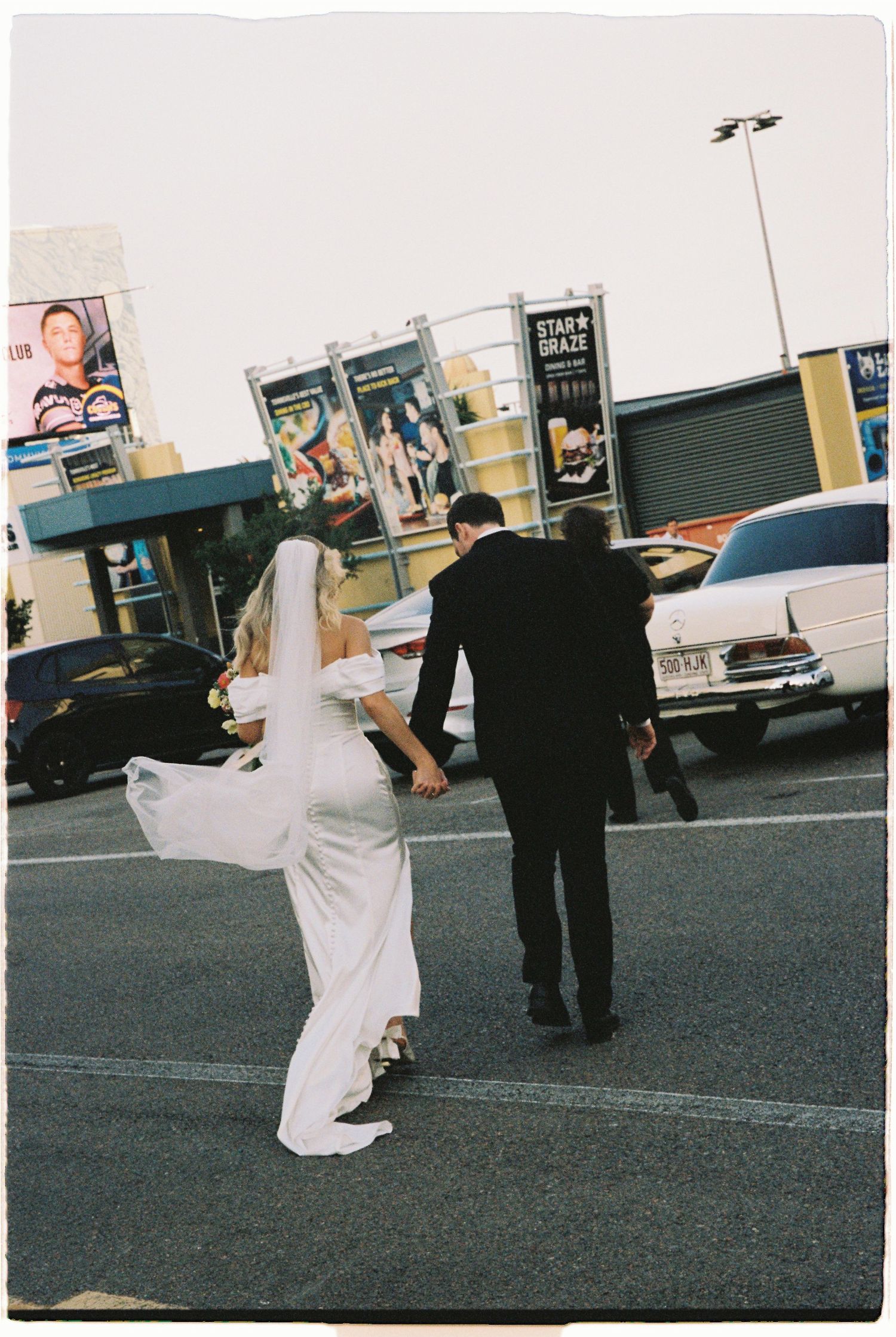 Bride and groom hold hands walking through parking lot, brides veil flowing behind her.
