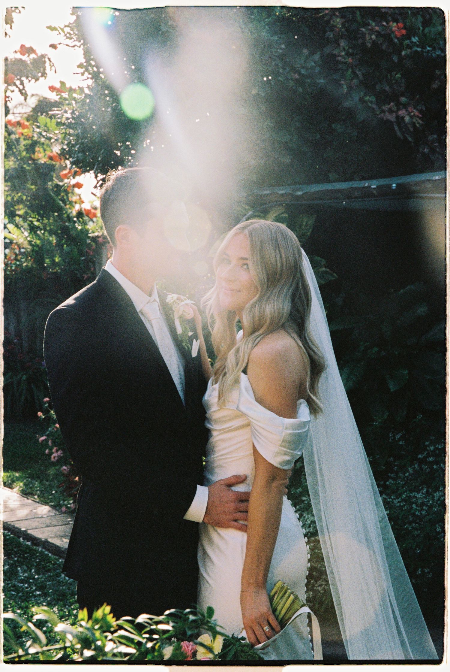 Bride in off-shoulder gown and veil embraces groom in black tuxedo in golden sunlit garden.