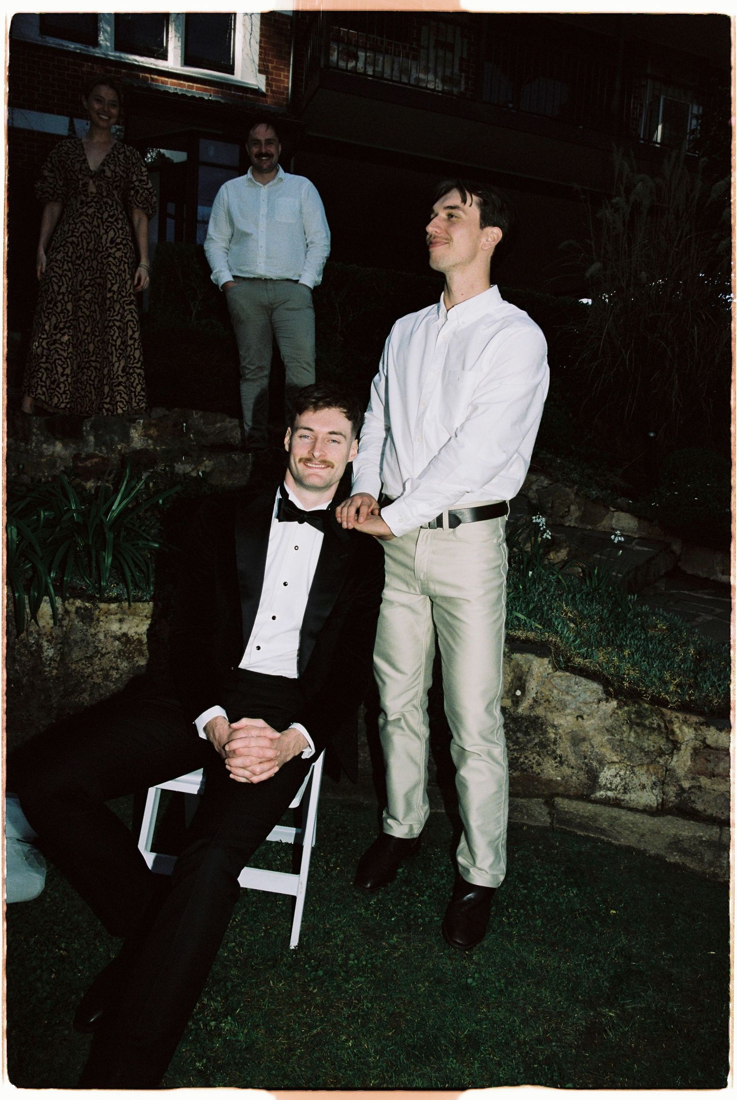 Two men pose at an outdoor wedding reception, one seated in a tuxedo and one standing in white shirt and khakis.
