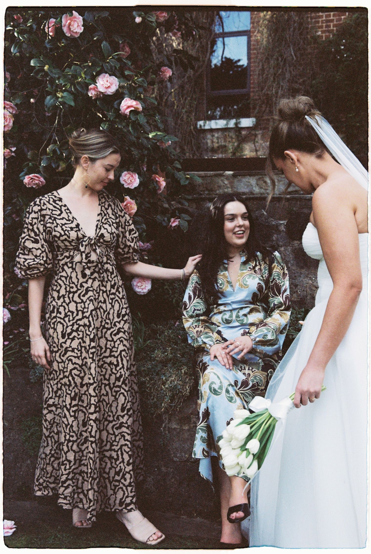 A bride holding white tulips chats with two guests in floral dresses beside a rose-covered stone wall.