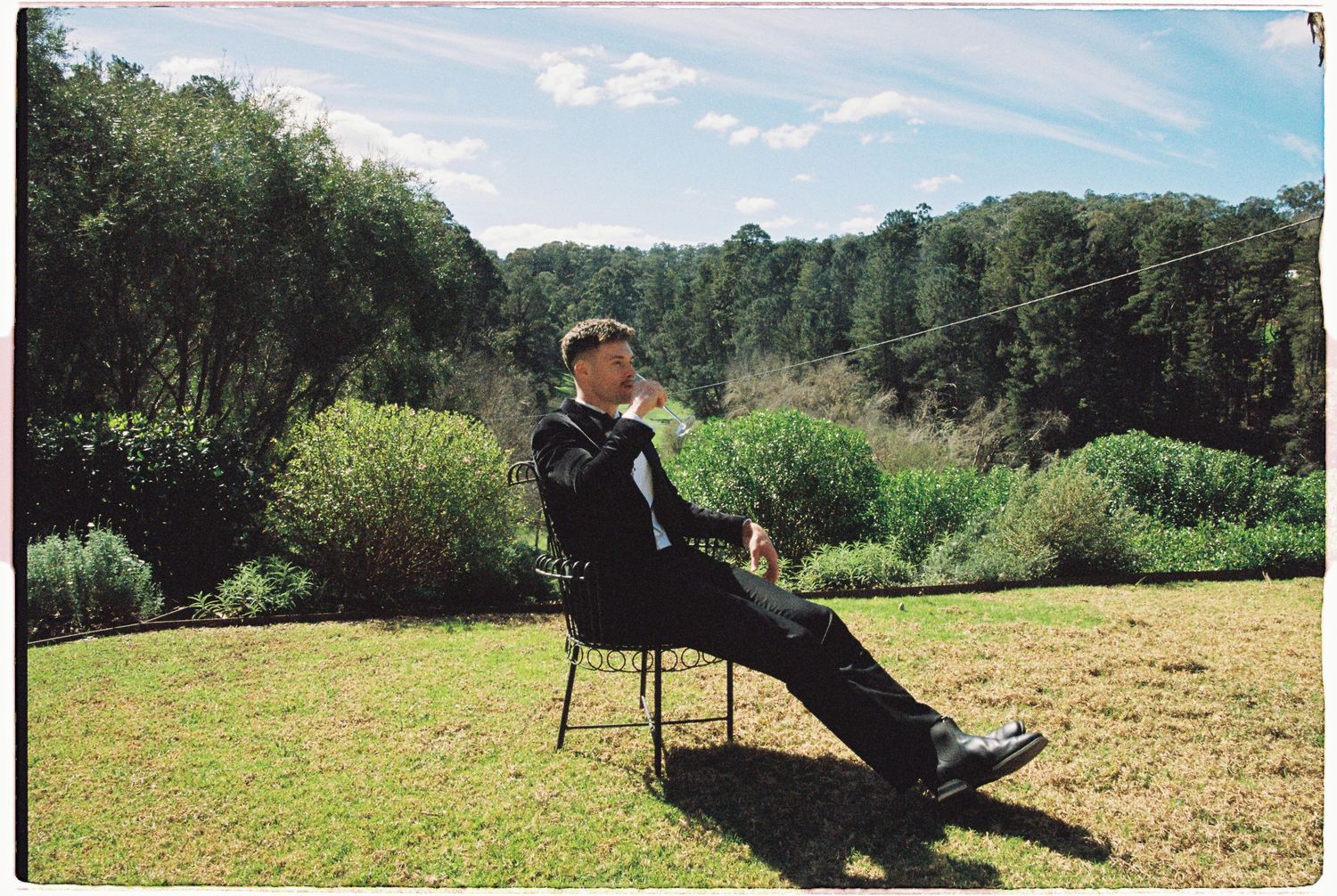 A man in a black suit relaxes on a chair in a sunny garden with trees and greenery in the background.