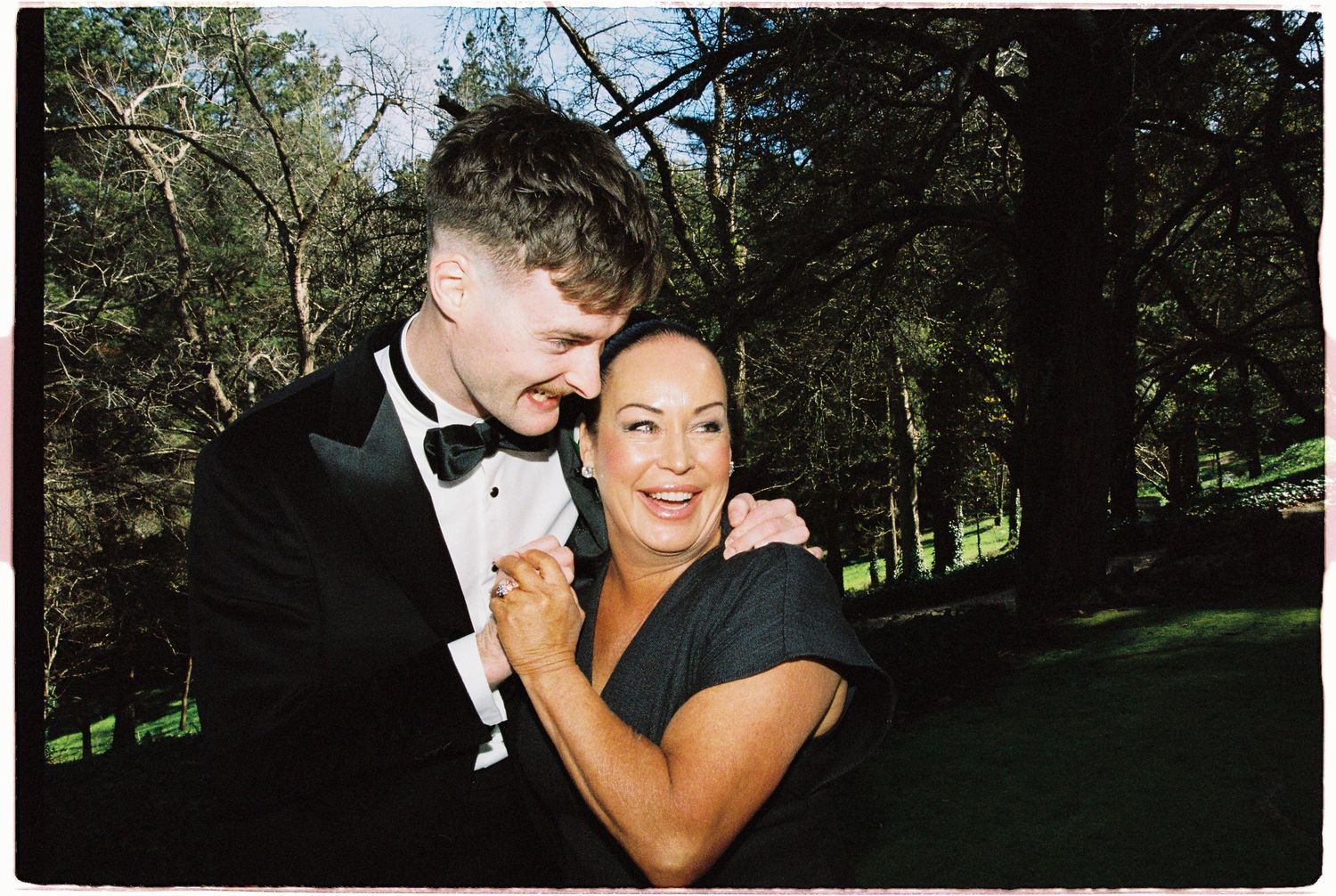 Groom in black tuxedo embraces smiling woman in black dress outdoors among tall pine trees.