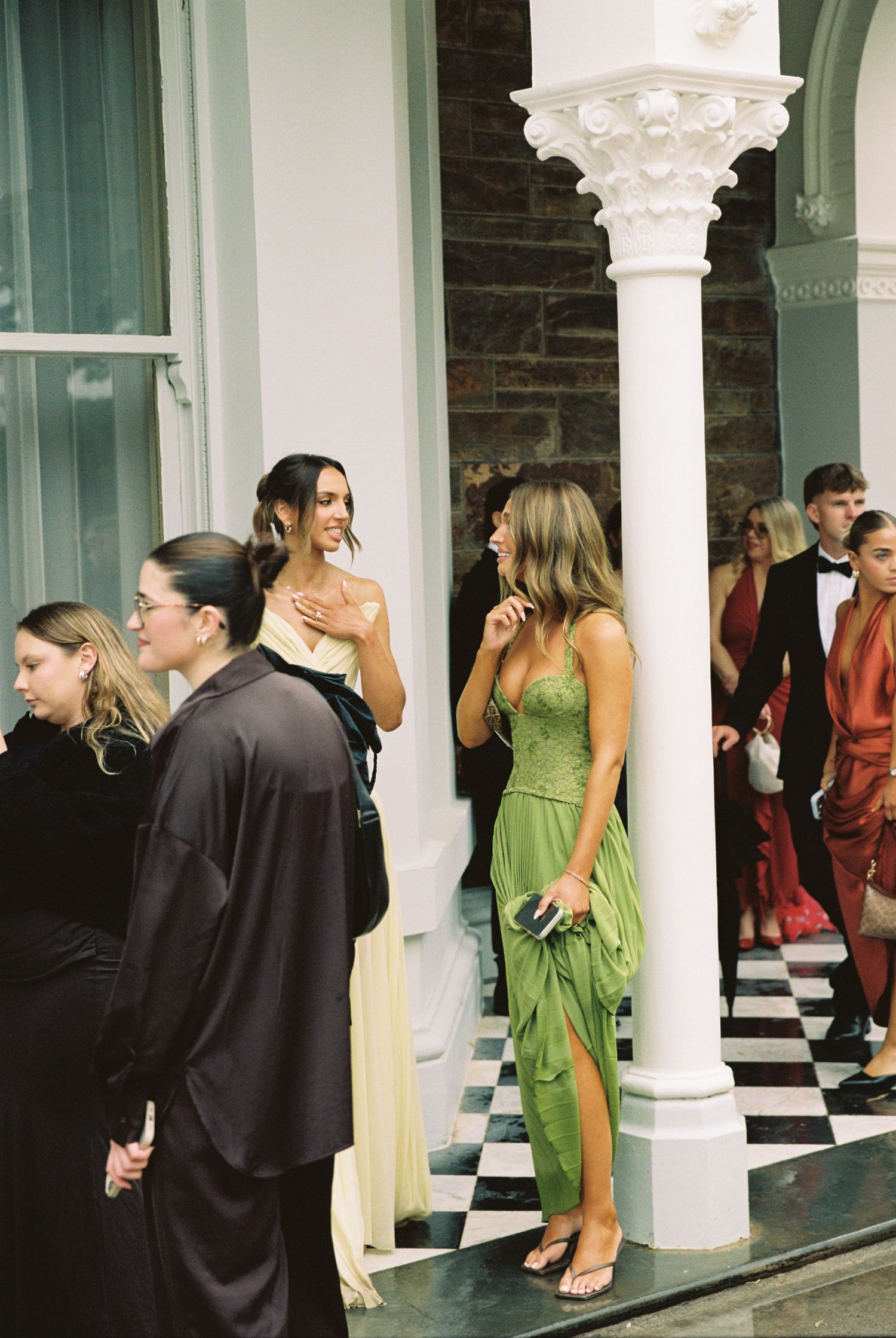 Elegantly dressed wedding guests mingle outside historic venue with white columns.