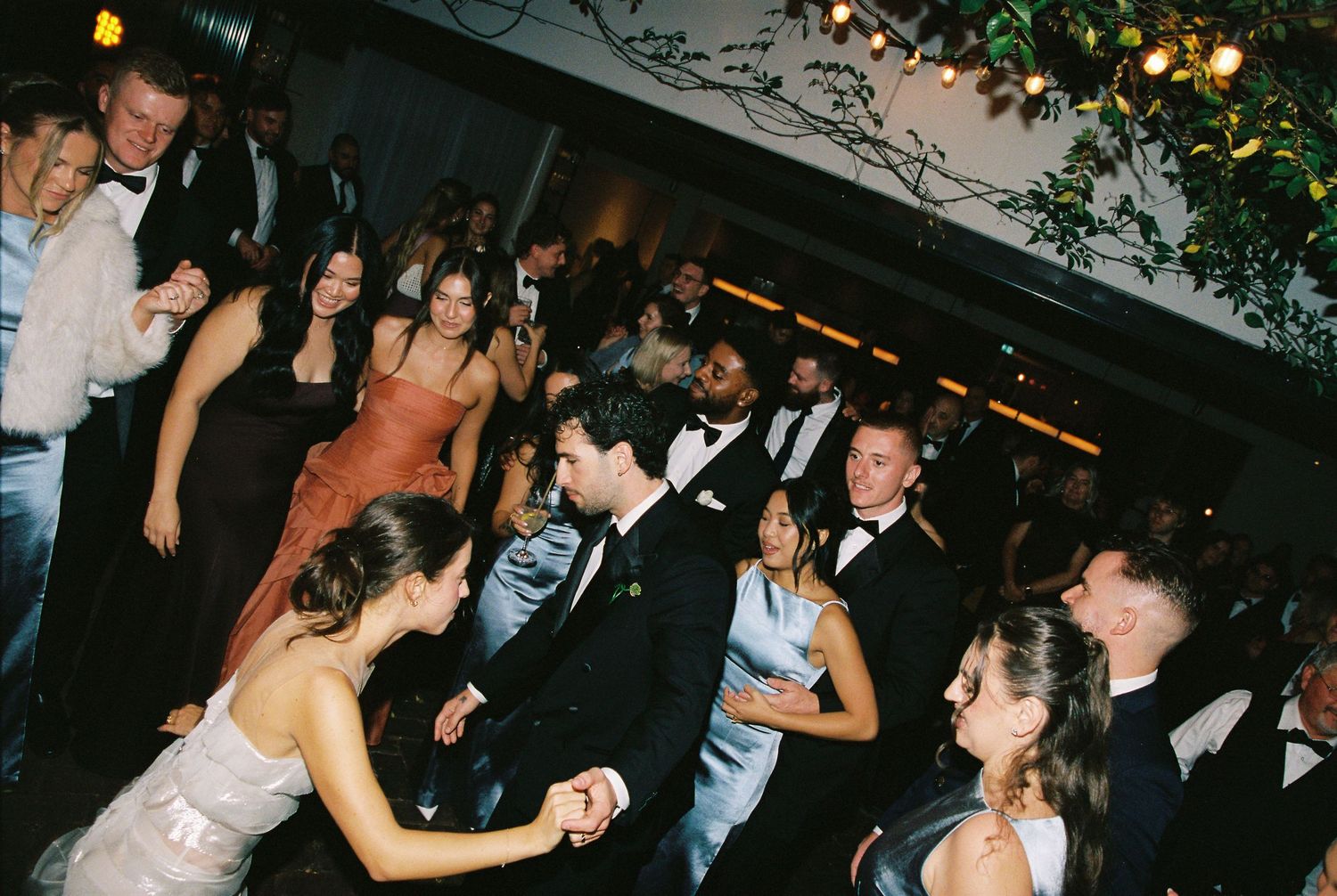 Guests in formal attire dance and mingle on a crowded wedding reception dance floor.