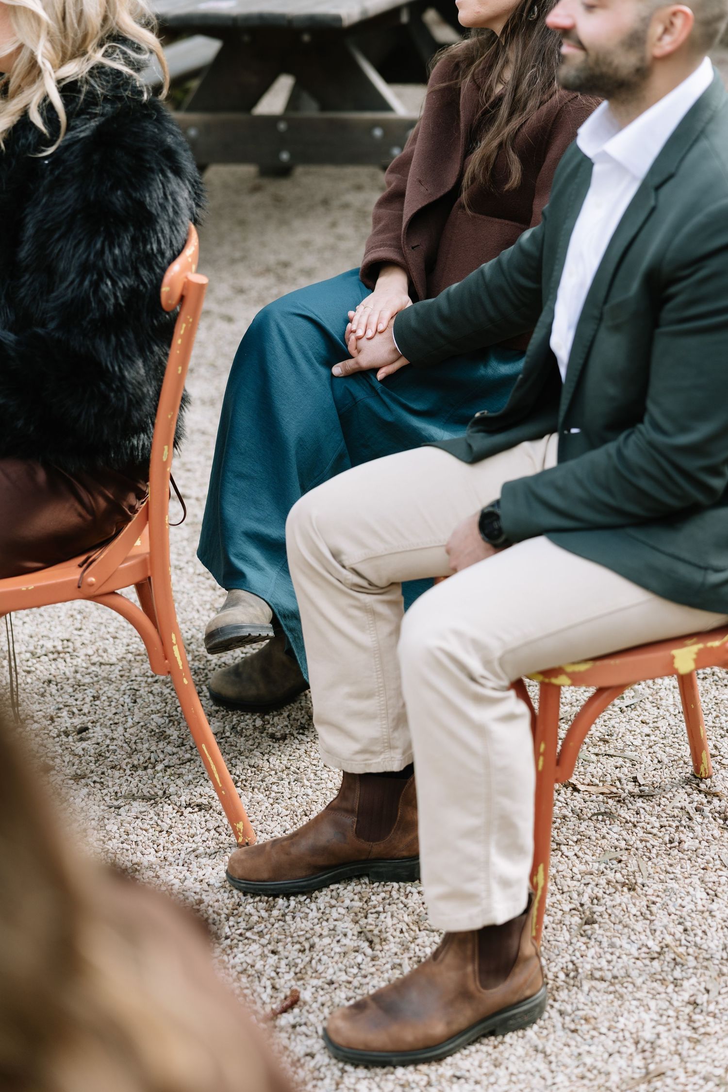 Couple holding hands while seated on orange chairs outdoors, dressed in elegant casual attire at a gathering.