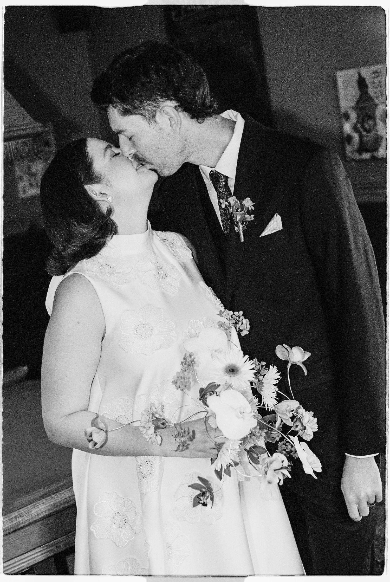 Black and white photo of a bride and groom sharing a kiss, bride holding a floral bouquet in a white dress.