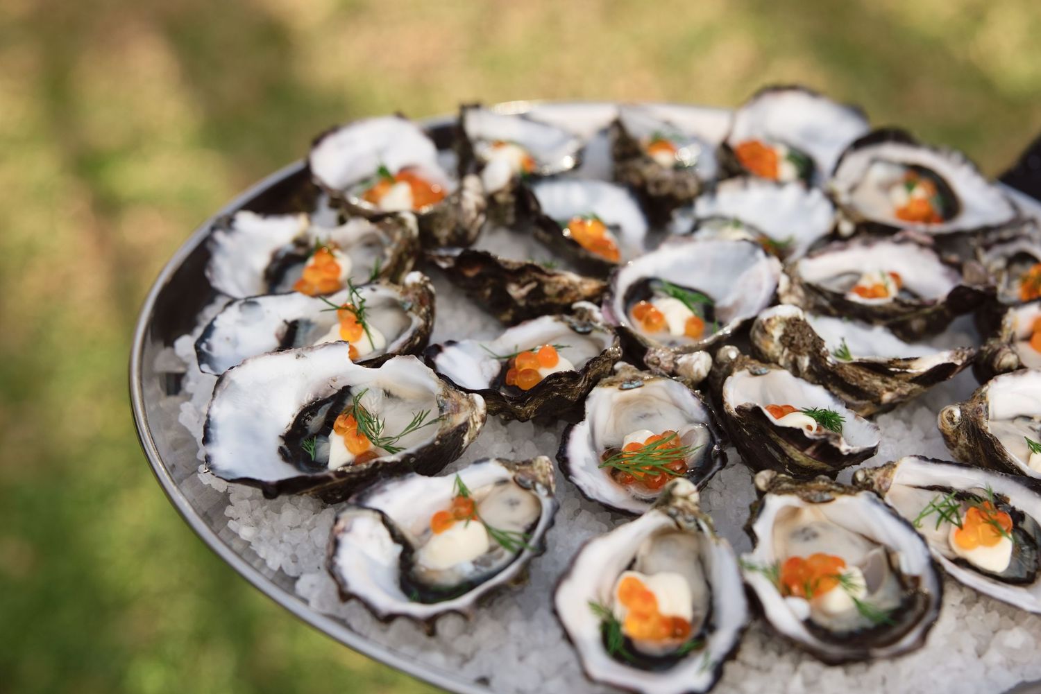 Fresh oysters on the half shell served on ice with garnishes on a silver platter outdoors.