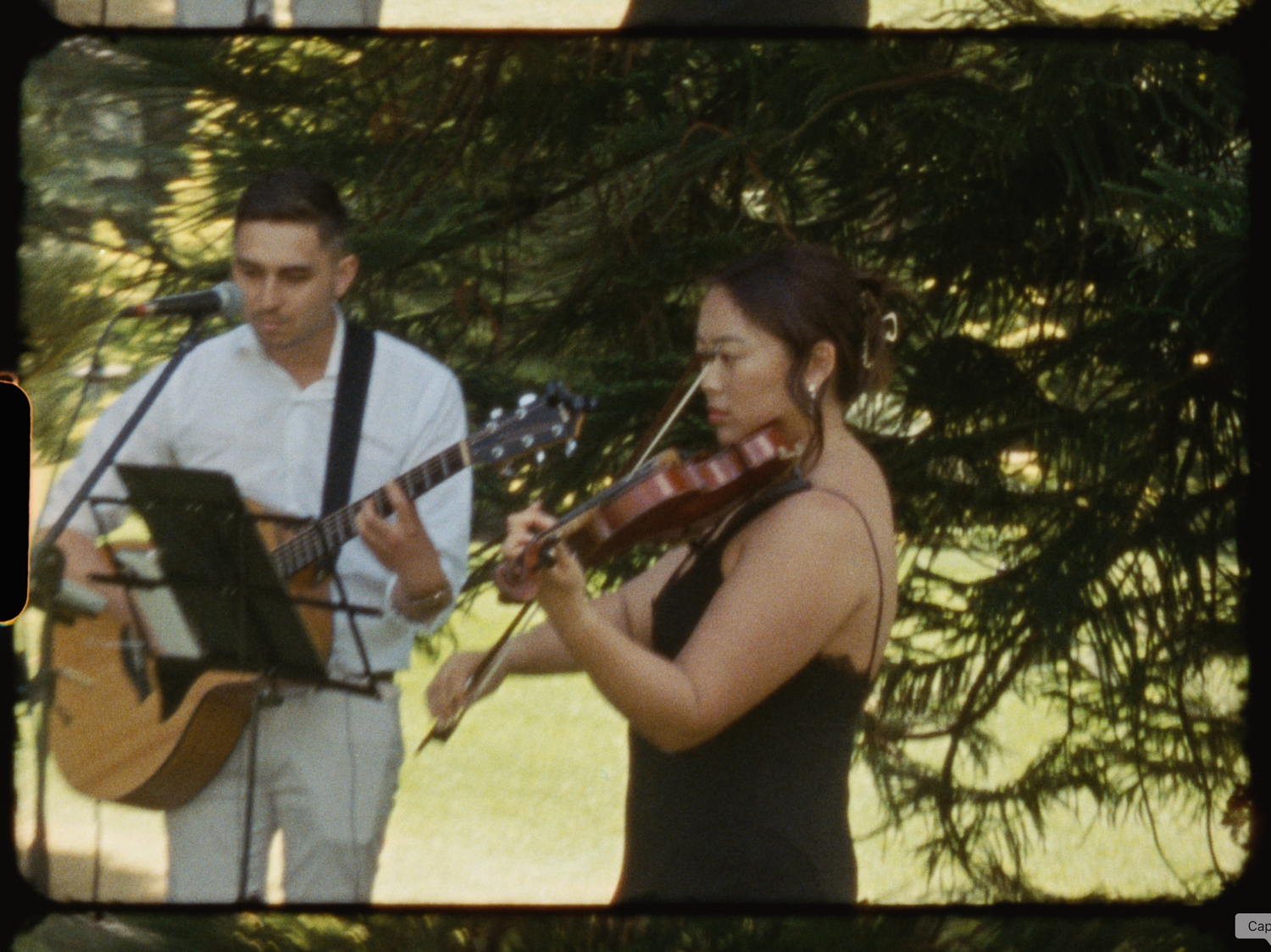 A man playing acoustic guitar and a woman playing violin perform outdoors near evergreen trees.