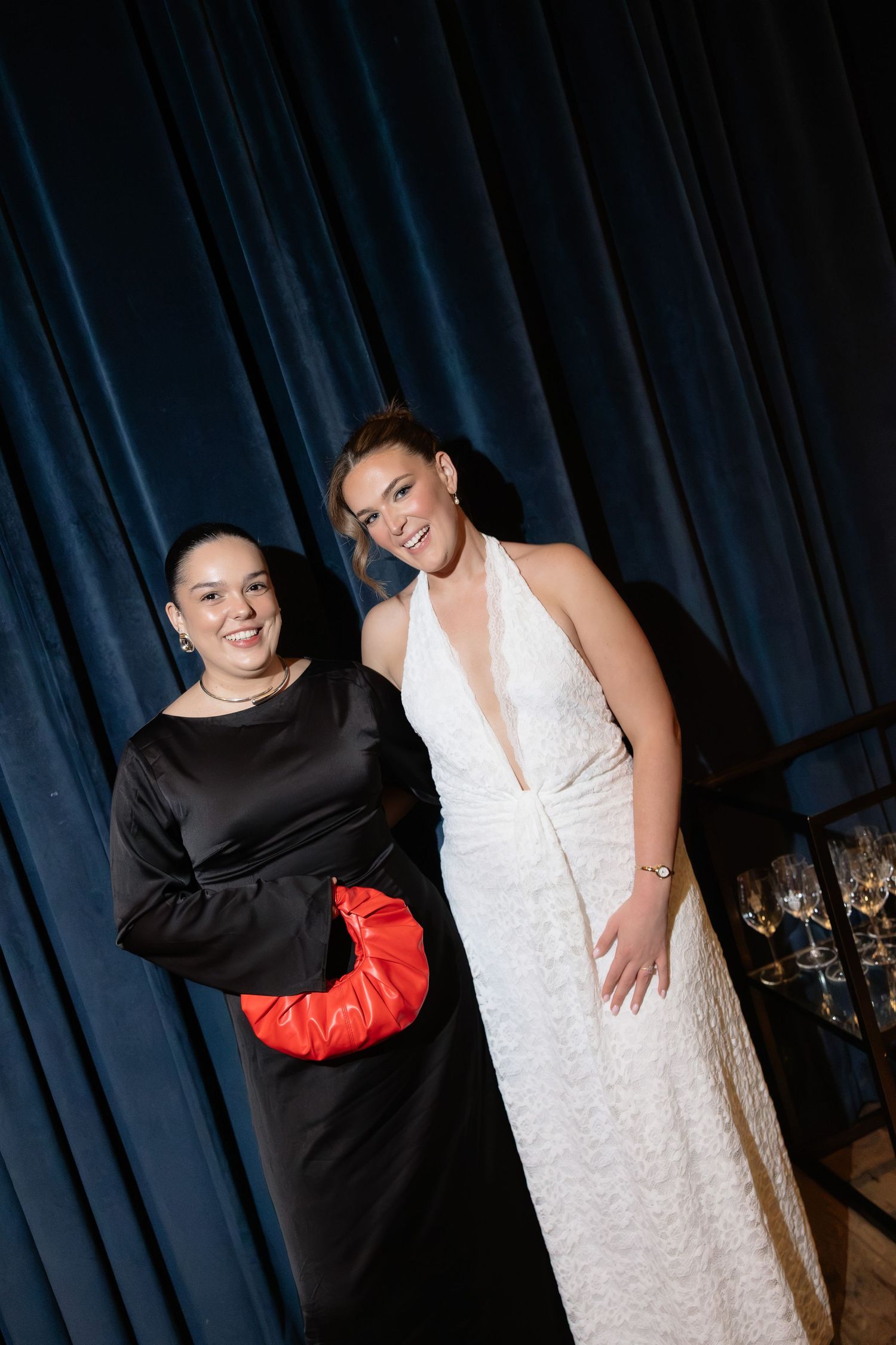 Two elegantly dressed women pose together at a formal event, one in black satin and one in a white halter gown.