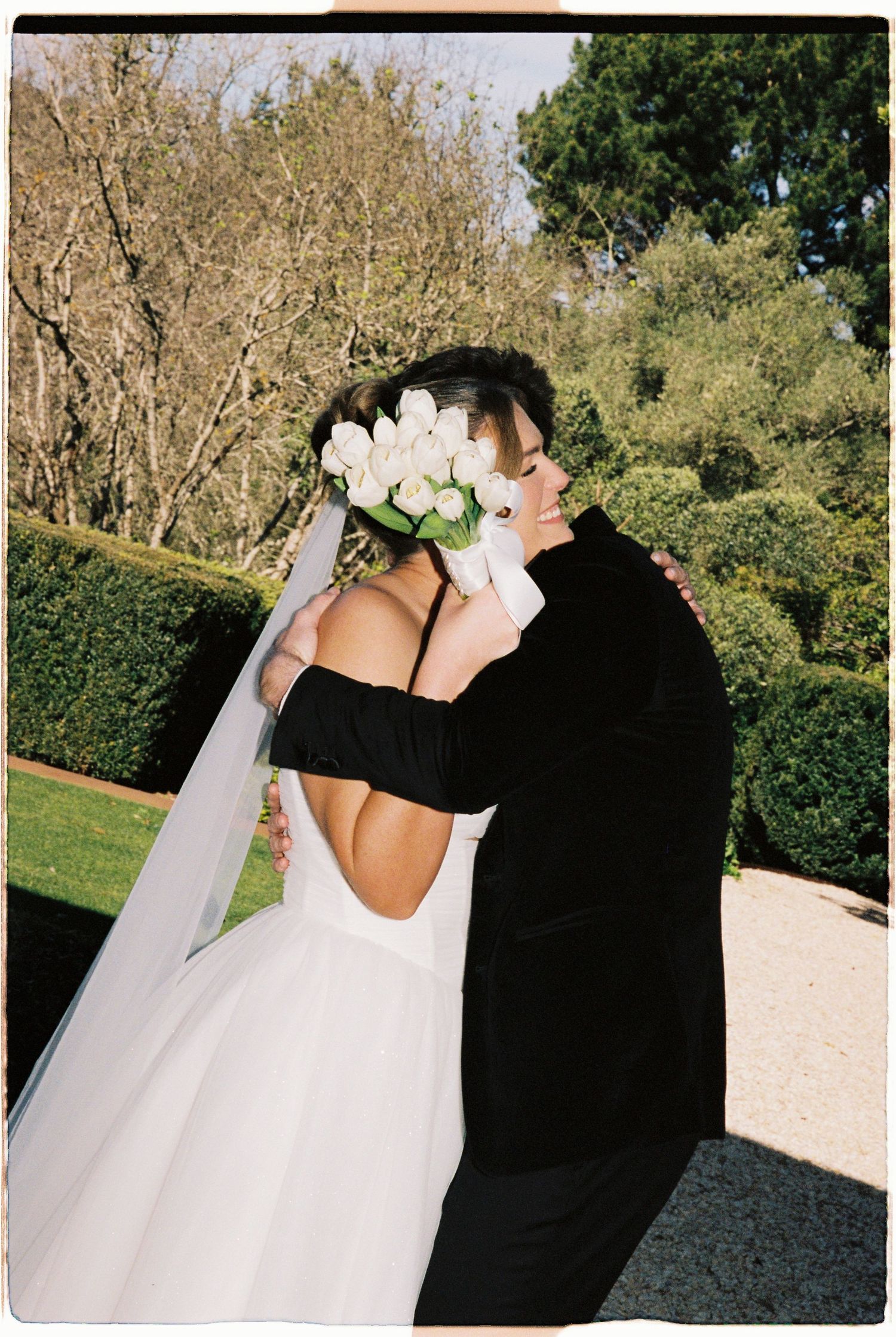 Bride in black off-shoulder gown with white tulip flower crown and veil embracing groom in black suit outdoors.