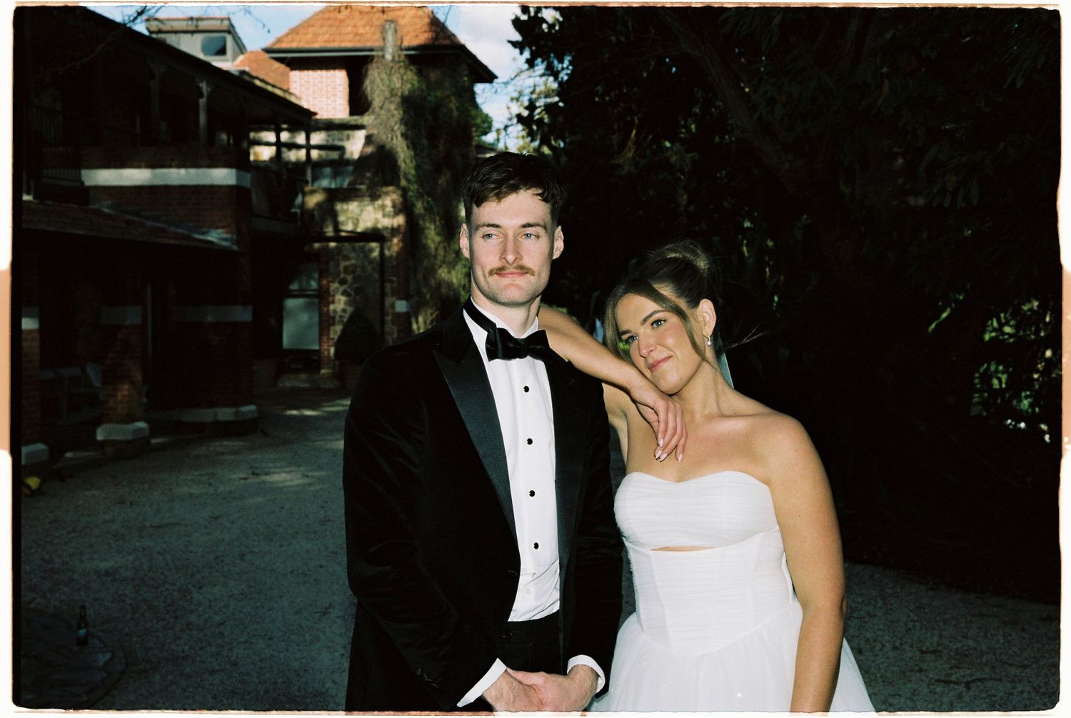 A couple in formal wedding attire, man in tuxedo and woman in white strapless gown, posing outdoors.