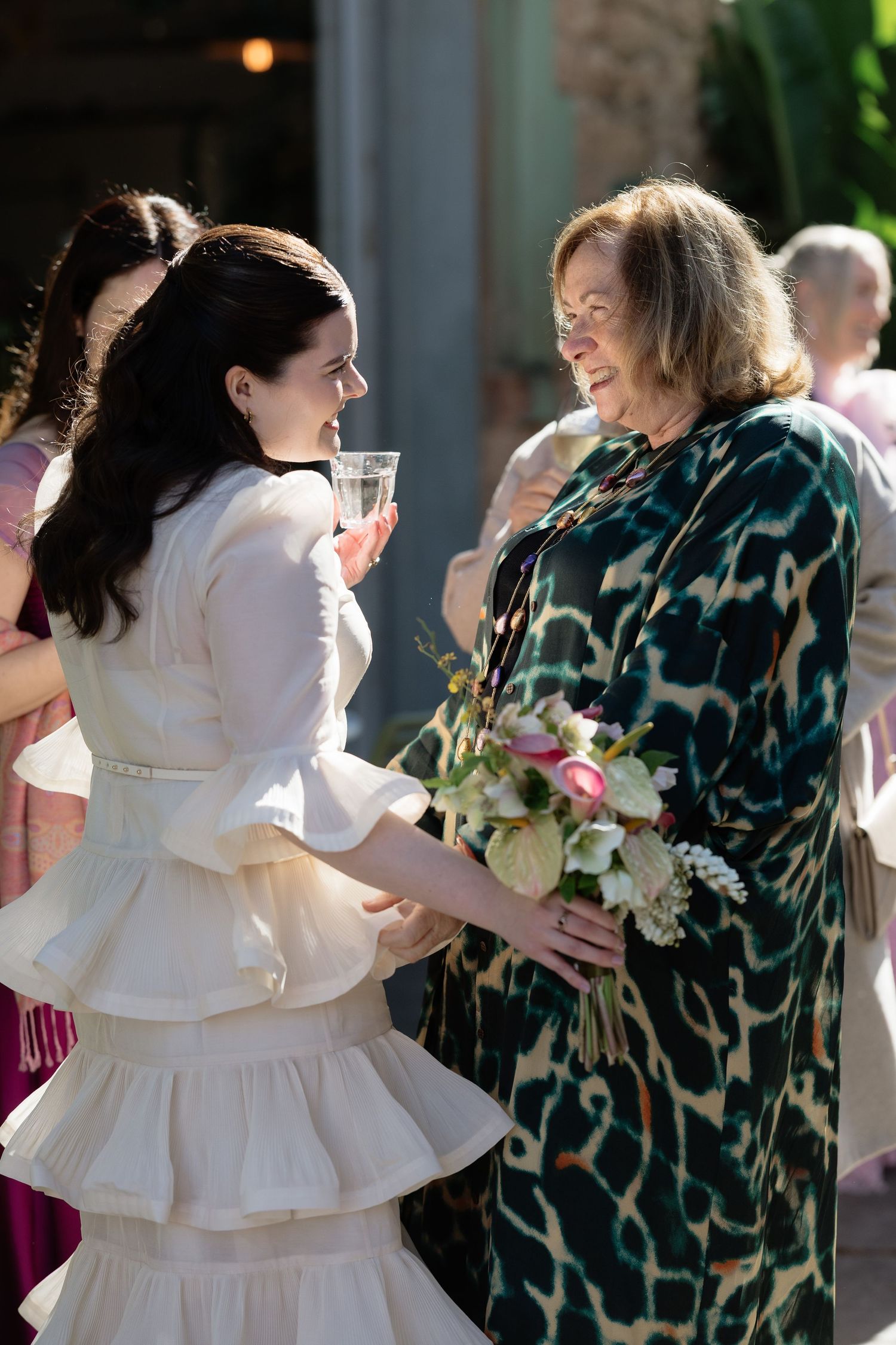 Bride in ruffled white dress smiling and chatting with older guest holding bouquet at wedding reception.