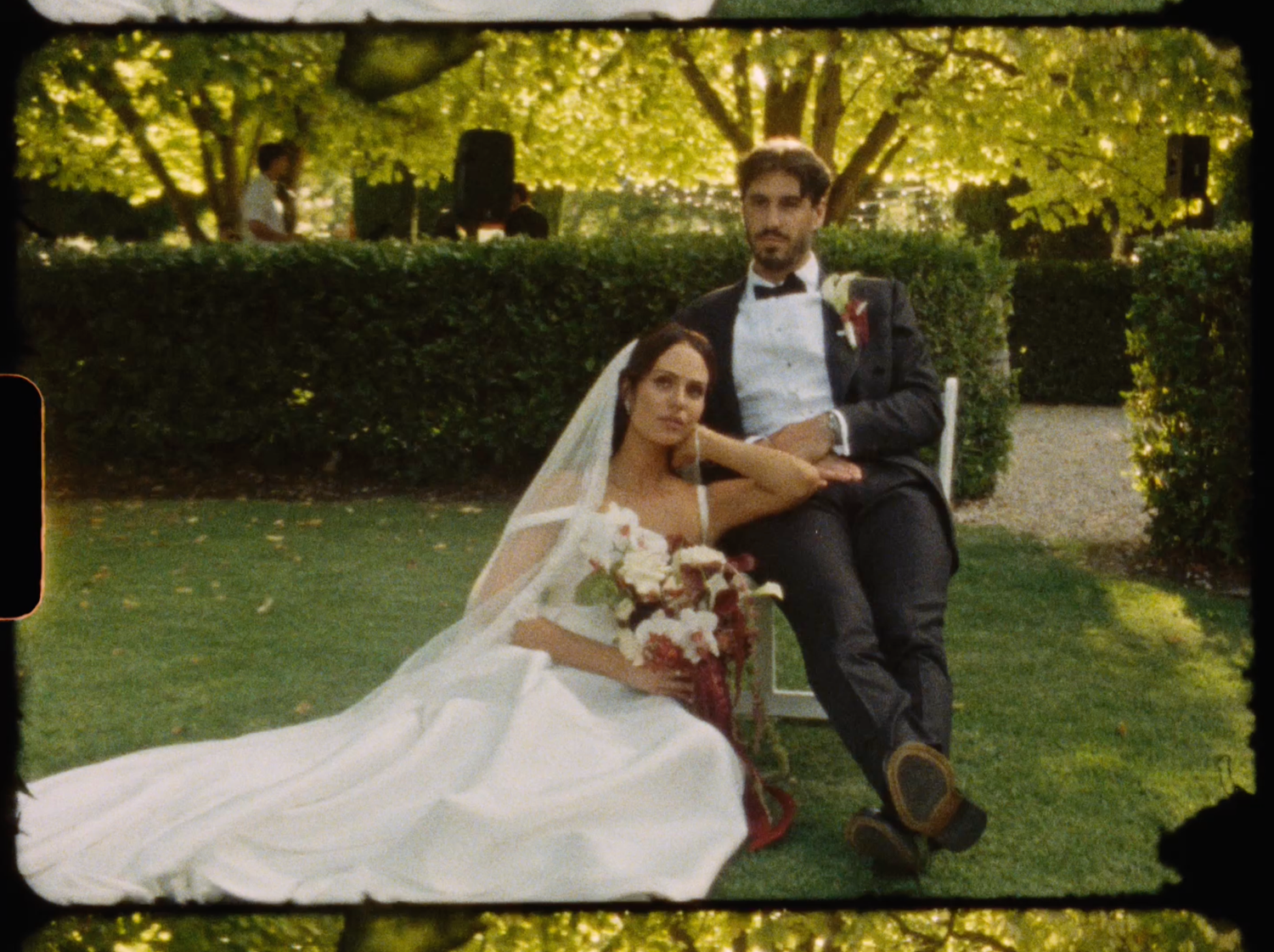 Bride in white gown and veil sits on grass as groom in tuxedo poses beside her in garden.