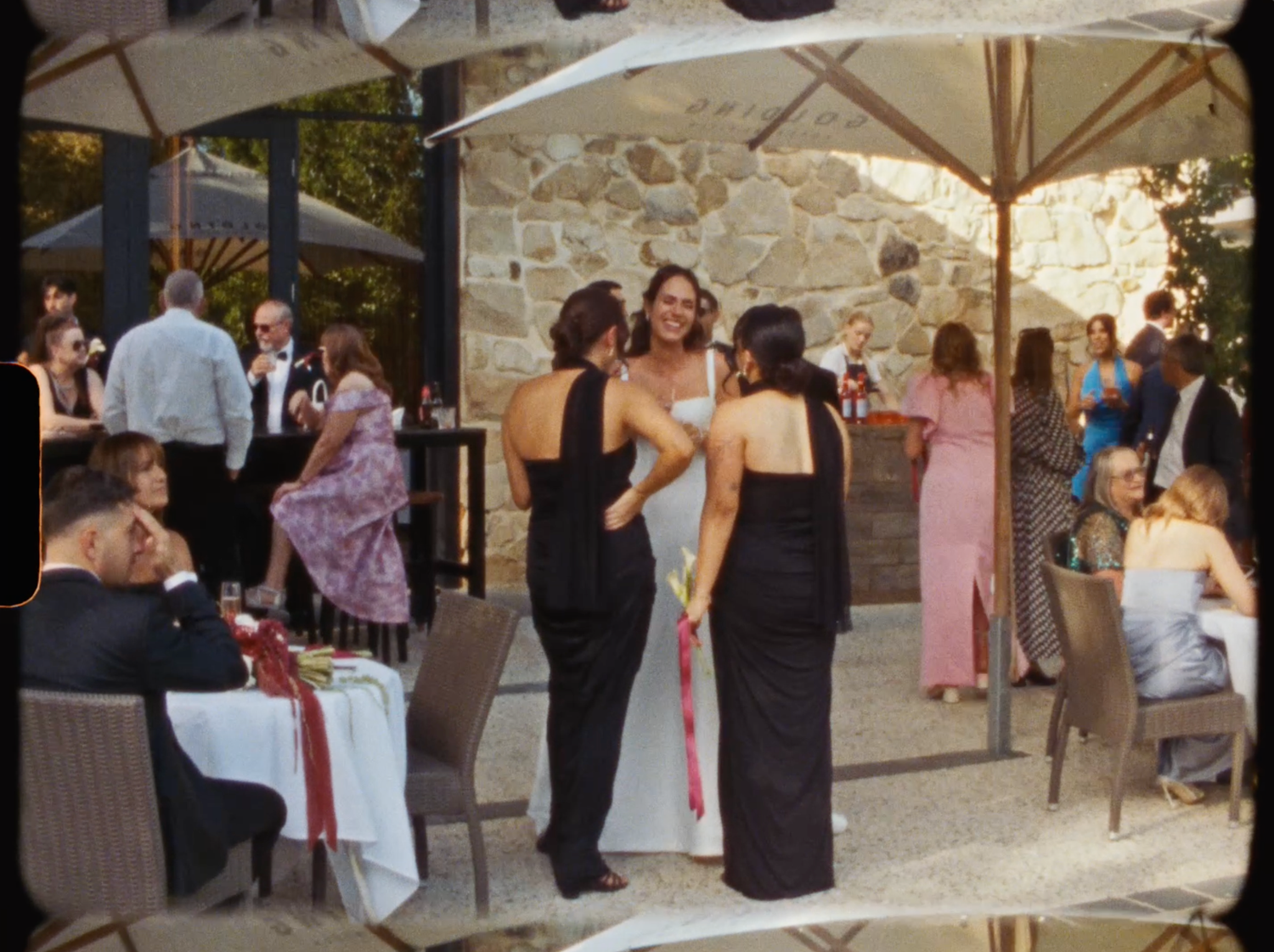 Wedding guests mingle outdoors at a stone venue, with bride in white gown surrounded by elegantly dressed attendees.