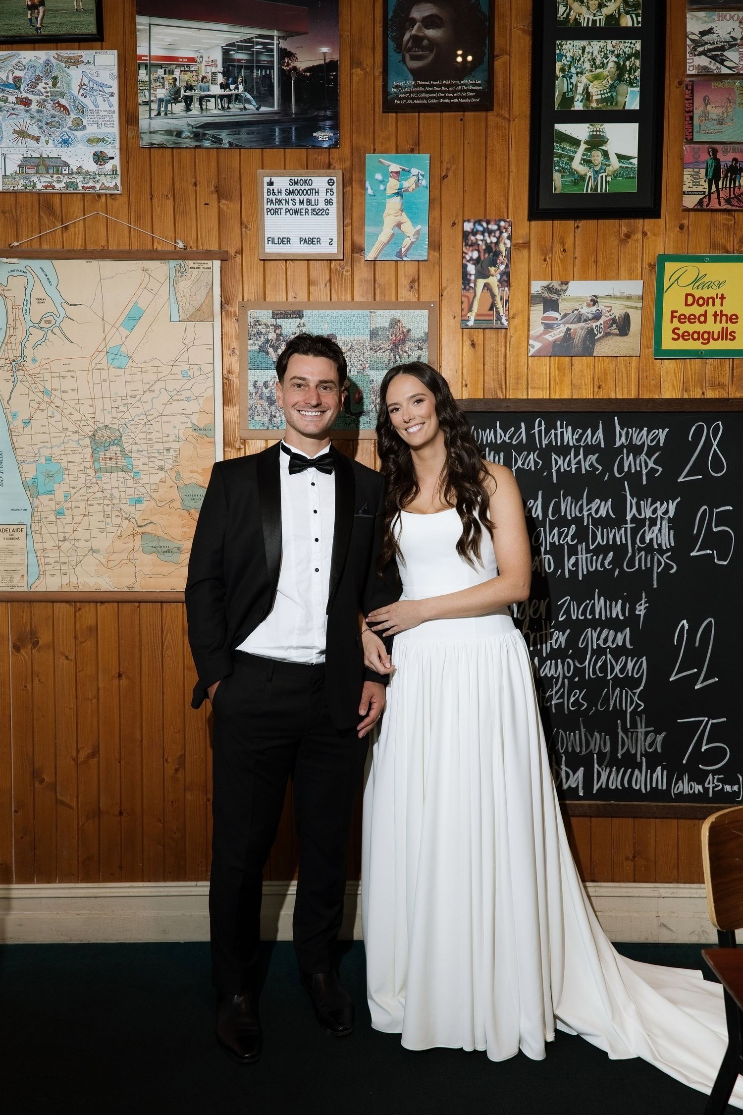 Wedding couple posing in front of a wooden wall with maps and a chalkboard menu in a pub.