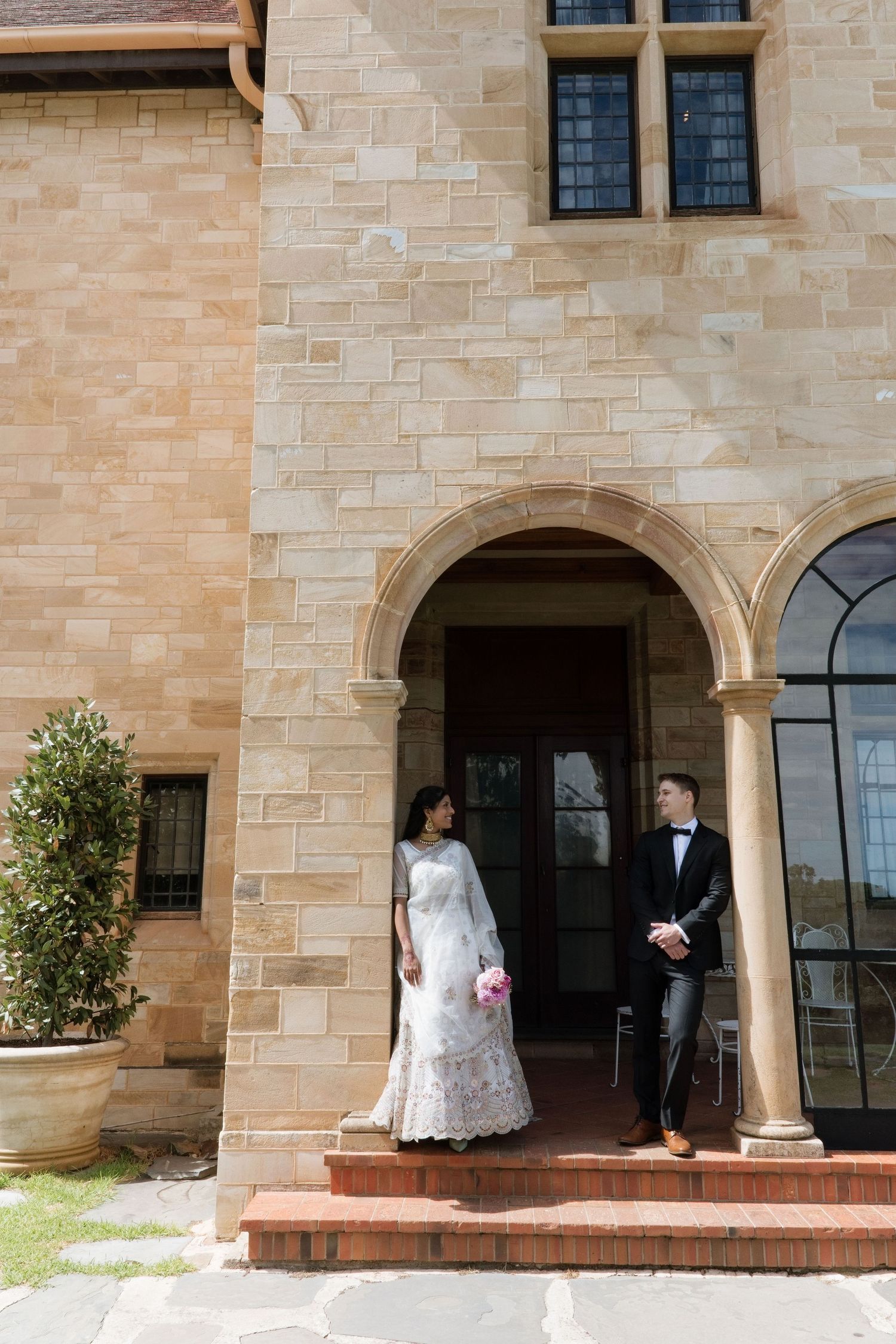 Bride and groom exchanging glances under ornate sandstone archway of historic building on their wedding day.
