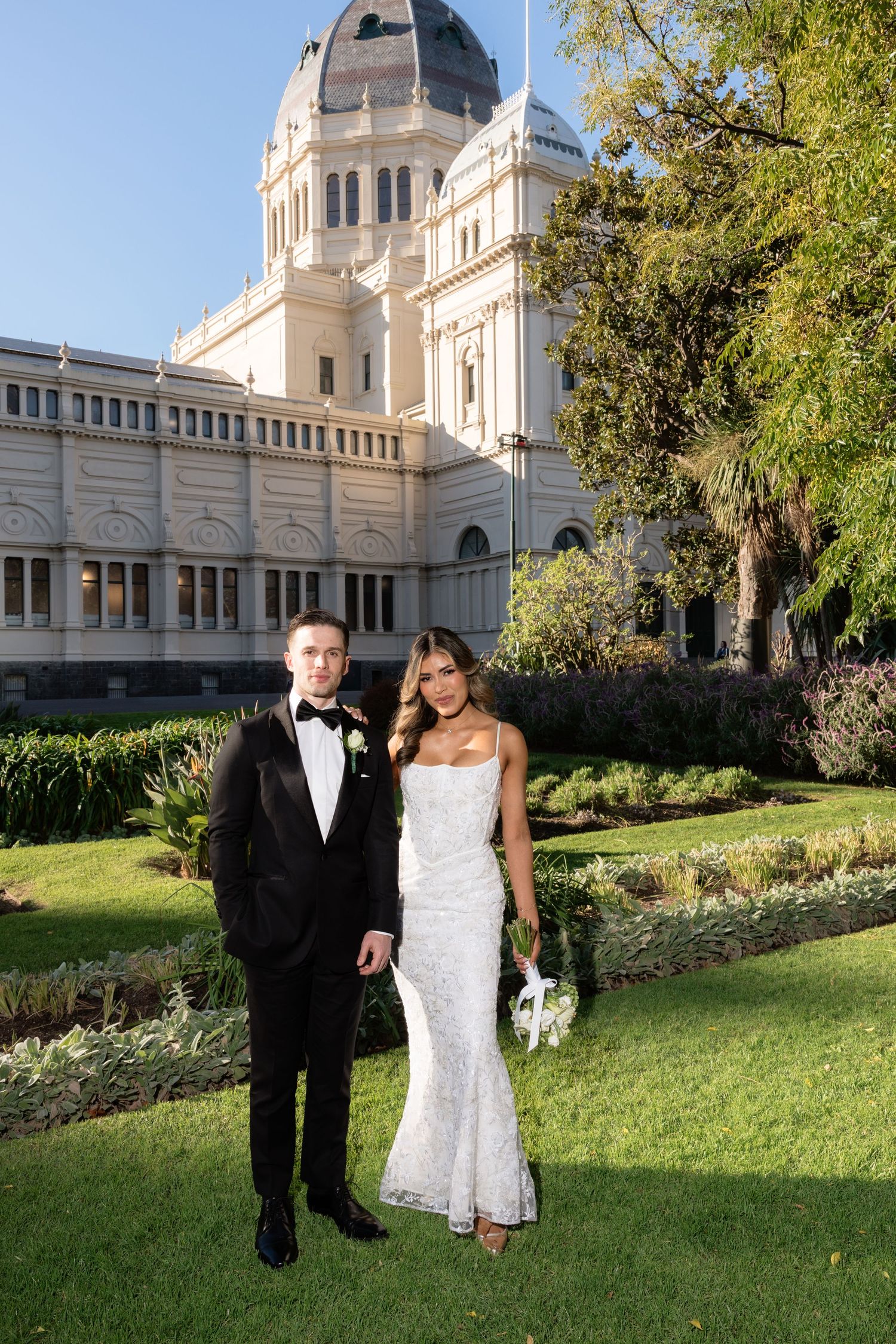 Elegant wedding couple posing in front of the Royal Exhibition Building in Melbourne, bride in white gown, groom in black tuxedo.
