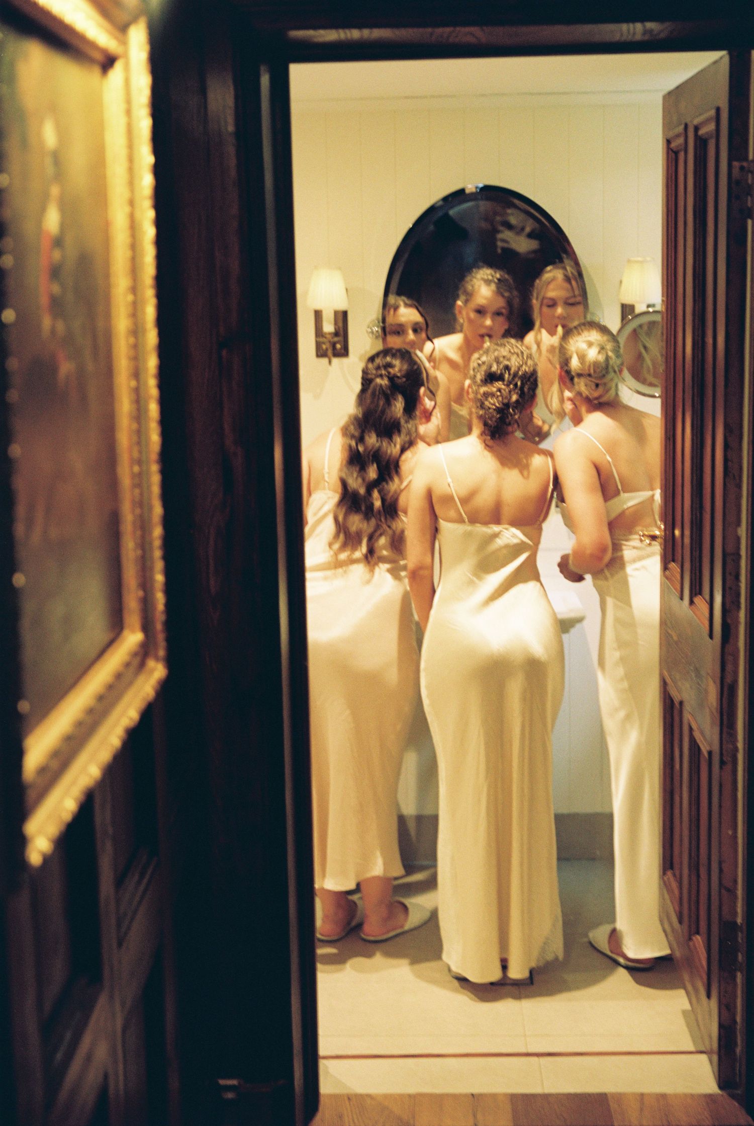 Bridesmaids in elegant ivory gowns gather around an oval mirror in a warmly lit room before a wedding ceremony.