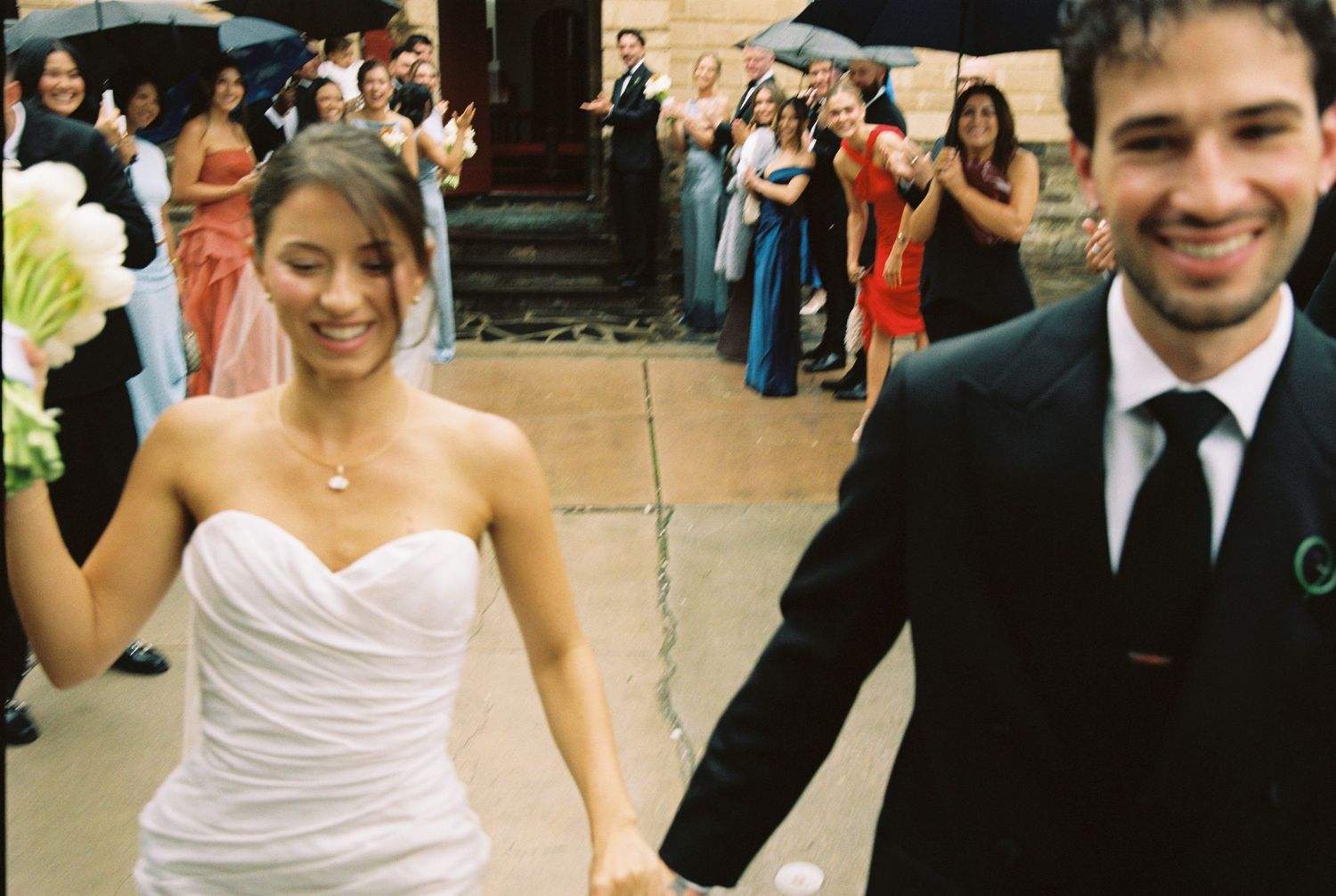 Smiling bride in white strapless gown and groom in black suit hold hands leaving wedding ceremony with guests cheering.