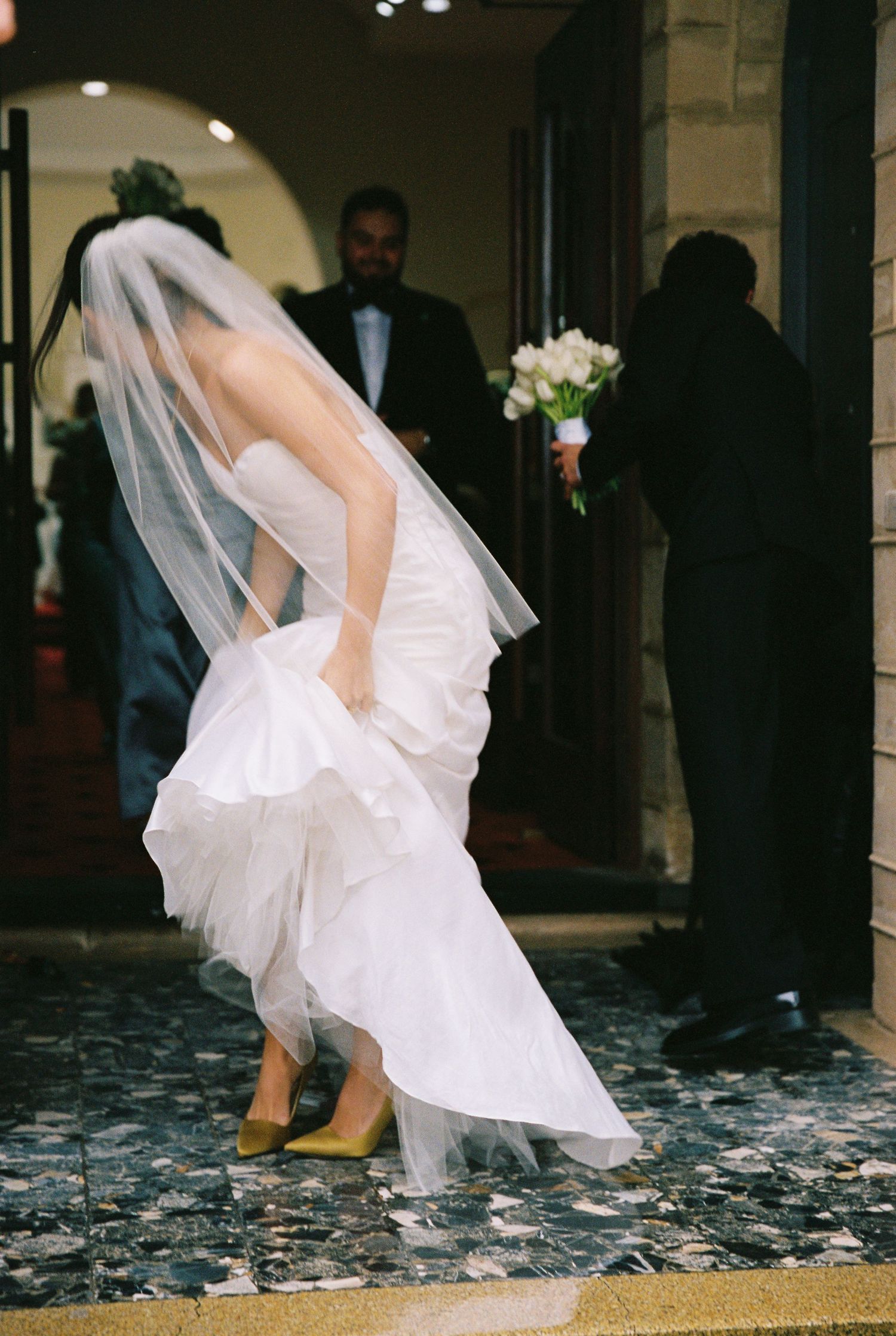 Bride in flowing white gown and veil with yellow heels exits a stone church doorway holding a bouquet.