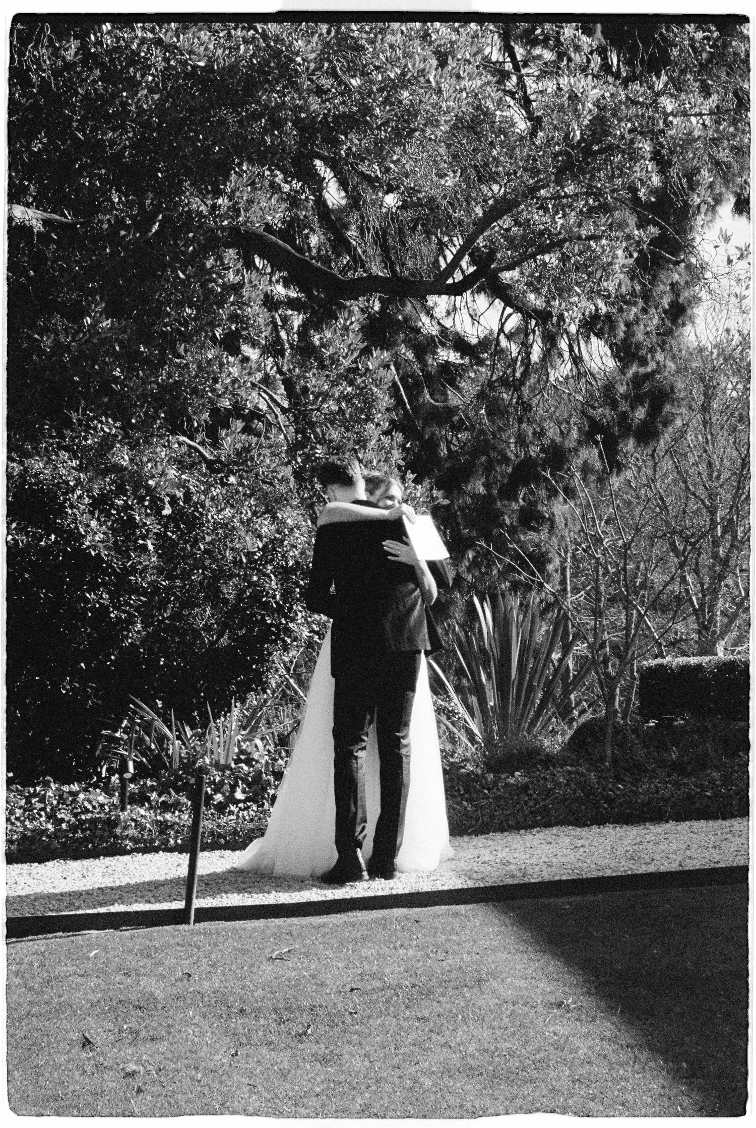 Black and white photo of a bride and groom embracing in a garden, viewed from behind, surrounded by lush trees.