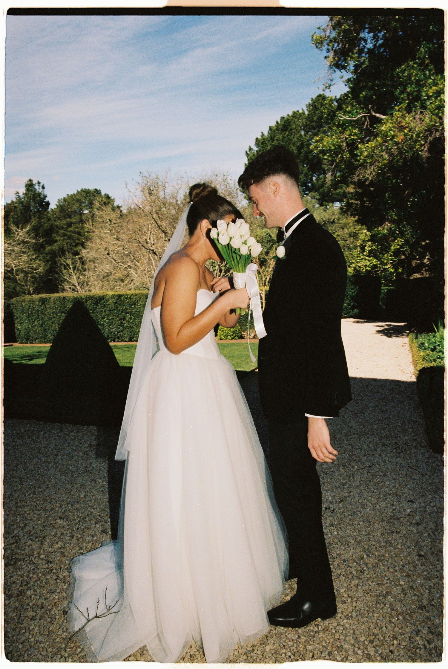 Bride and groom in formal attire share intimate moment with white tulip bouquet at elegant outdoor garden wedding.