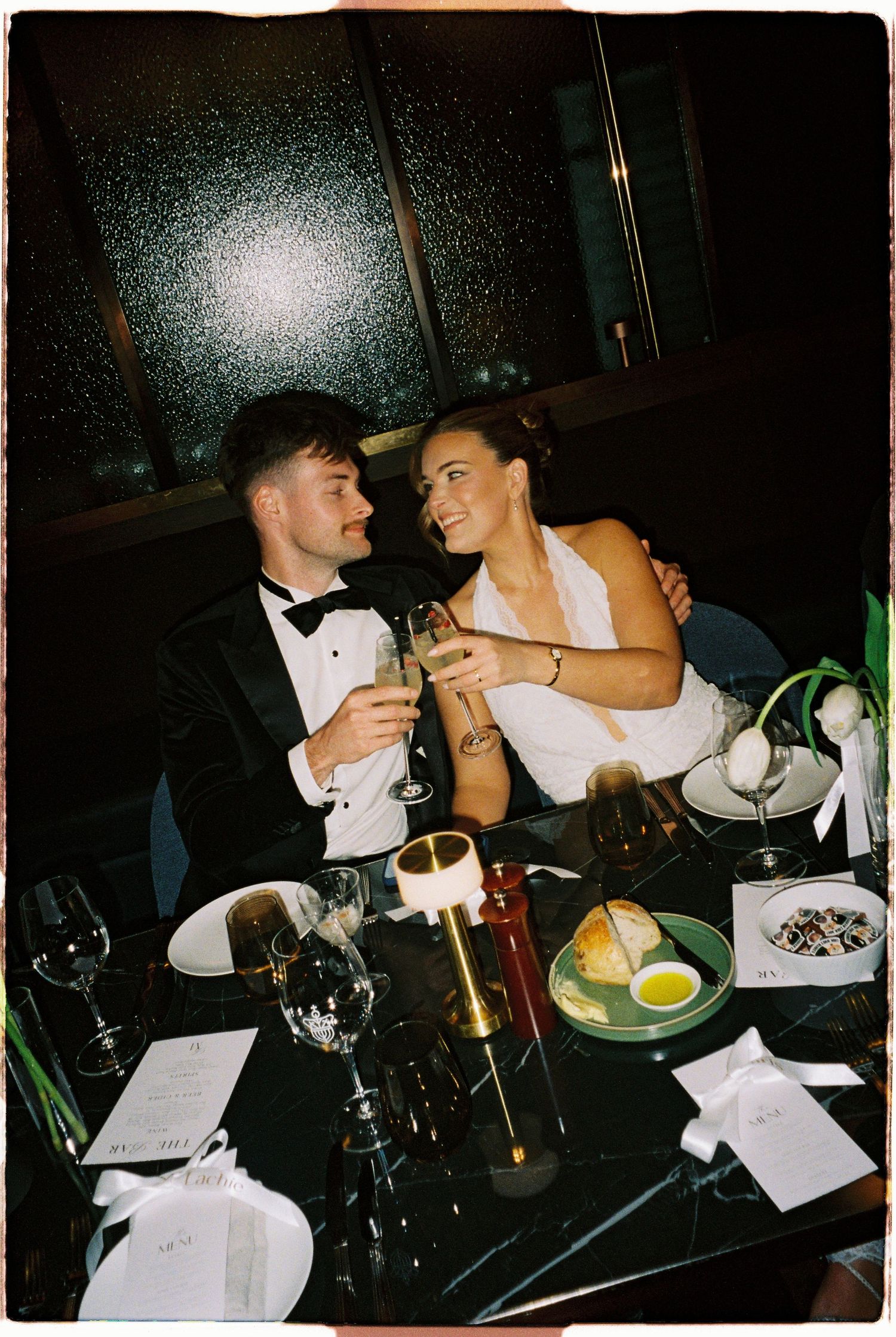 Newlyweds toast with champagne glasses at a candlelit wedding reception dinner table, smiling at each other.