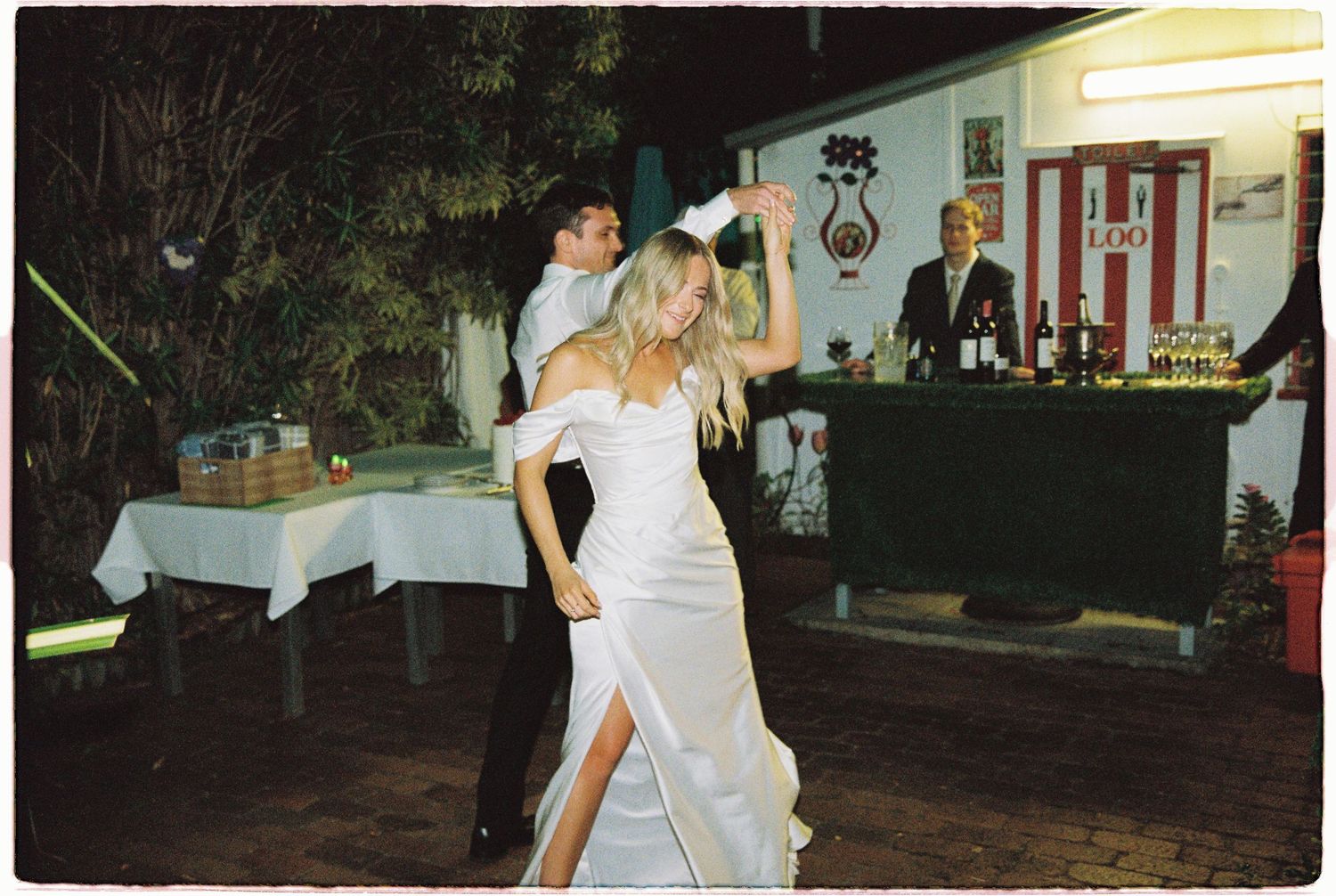 Bride and groom dancing energetically at outdoor evening wedding reception near decorated bar area.