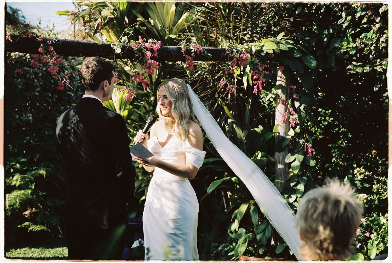 Bride in white off-shoulder gown reads vows at outdoor garden ceremony with lush tropical backdrop.
