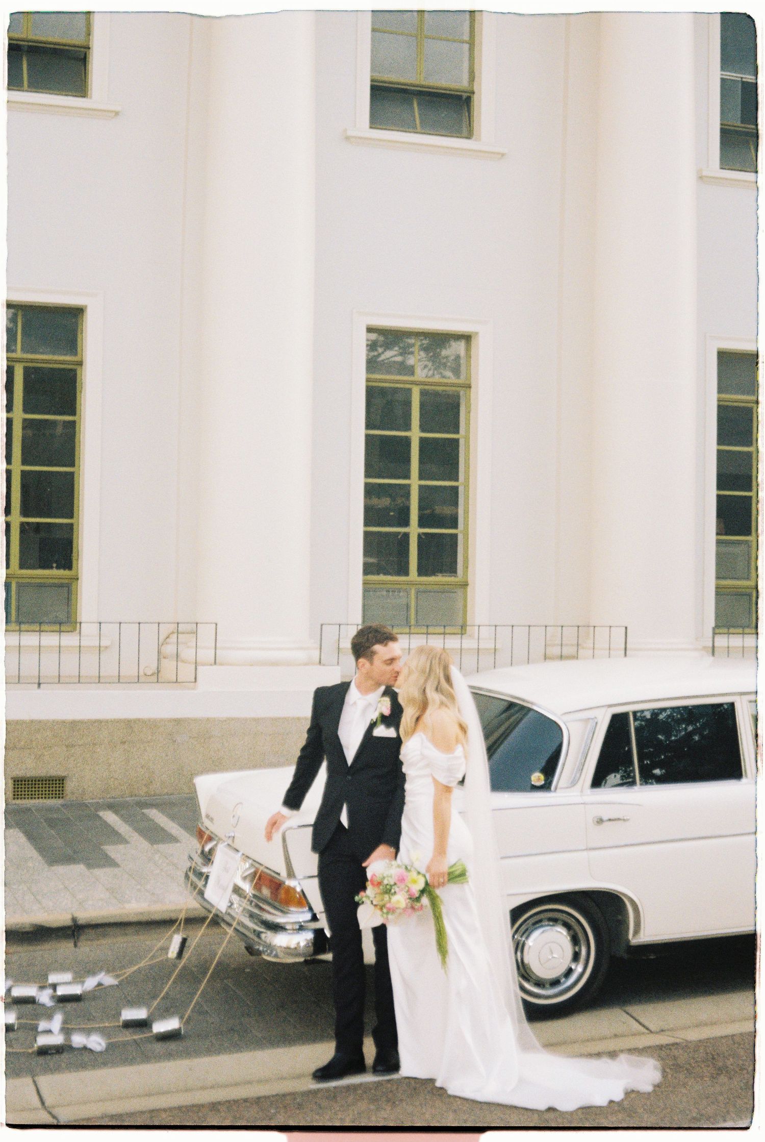 Newlyweds kiss beside vintage white classic car decorated with tin cans outside white columned wedding venue.