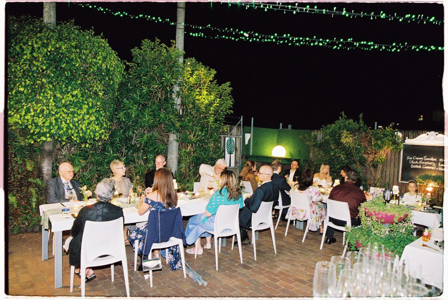 Guests dining outdoors at night under string lights surrounded by lush greenery at an elegant garden dinner party.