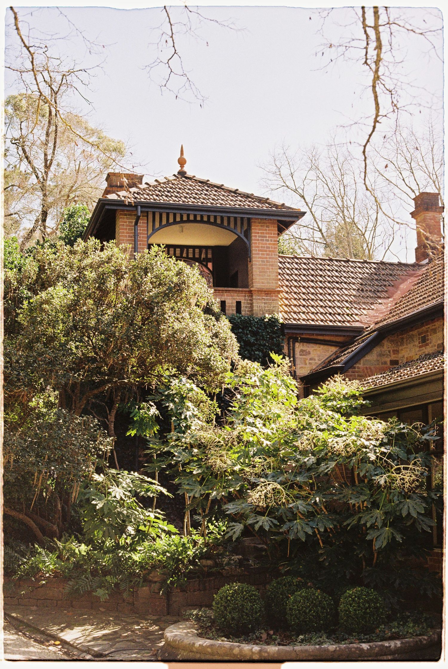 Historic brick home with tiled roof turret surrounded by mature trees and lush green garden foliage.