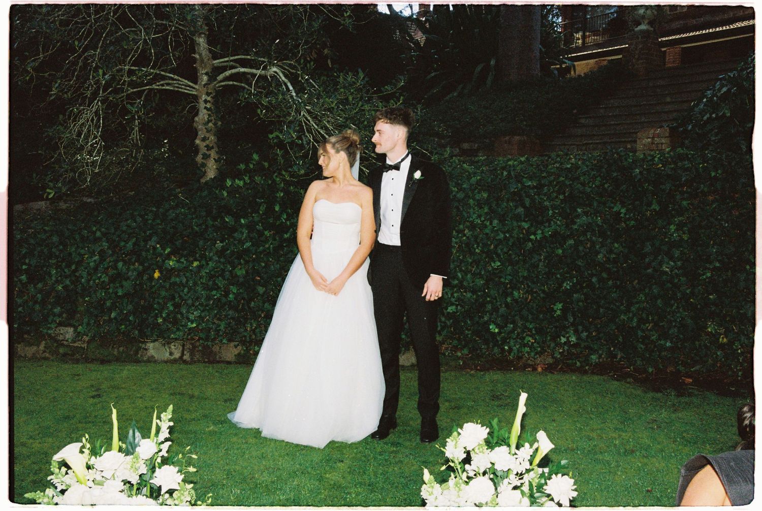 Bride and groom share a romantic moment at outdoor wedding ceremony surrounded by lush greenery and white floral arrangements.