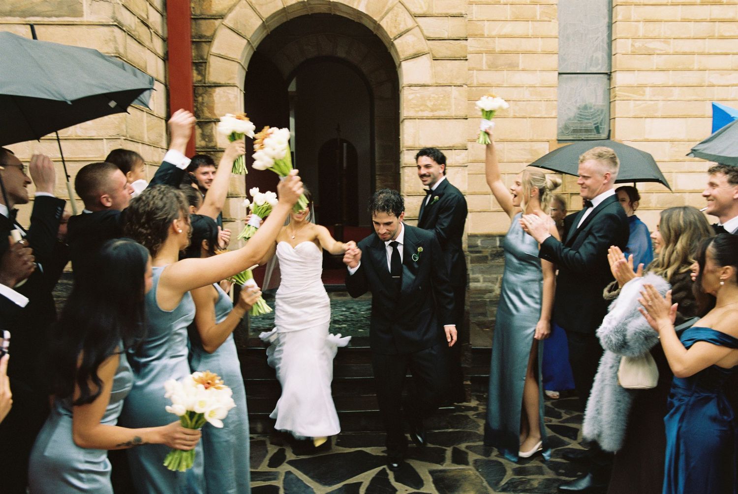 Bride and groom celebrate wedding exit through cheering guests holding bouquets outside historic stone building.