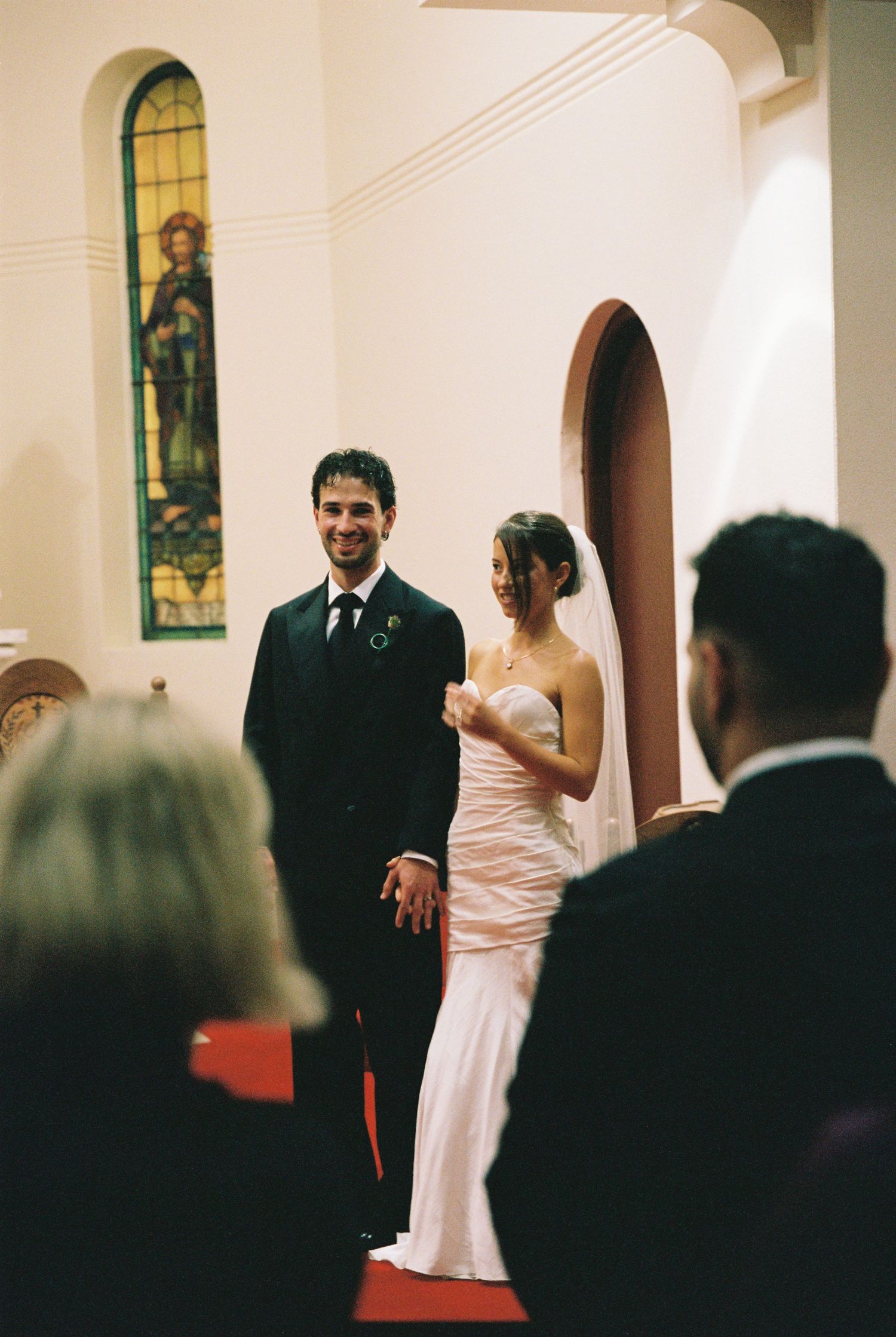 Smiling groom in black suit holds brides hand at church altar with stained glass window behind.