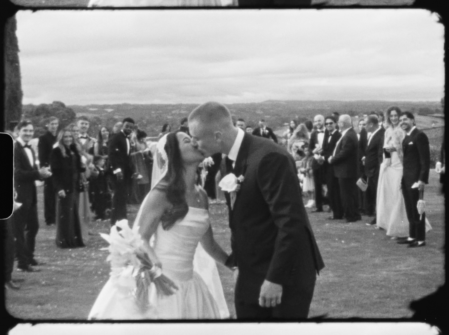 Black and white photo of a bride and groom sharing their first kiss at an outdoor wedding ceremony with guests applauding.