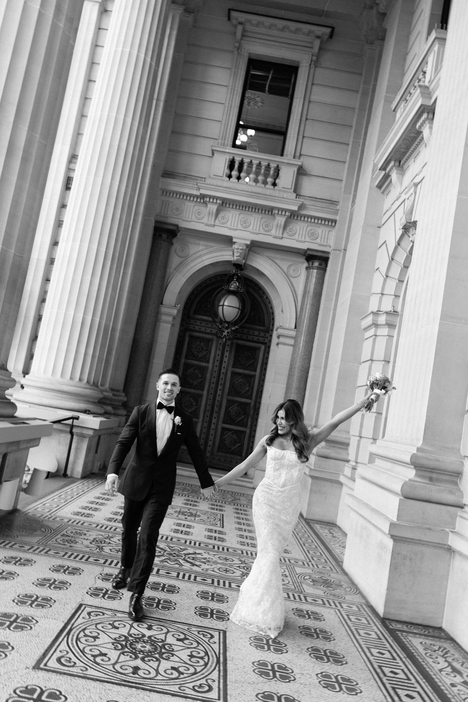 Bride and groom walk hand-in-hand past grand columns on ornate tiled steps in elegant black and white wedding photo.