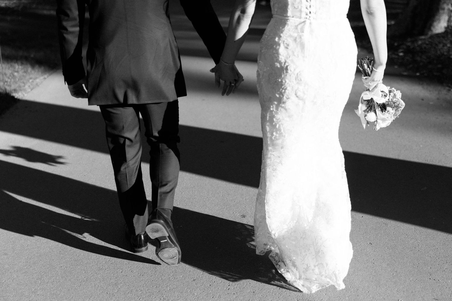 Black and white photo of bride and groom walking hand in hand, bride holding bouquet in elegant wedding attire.