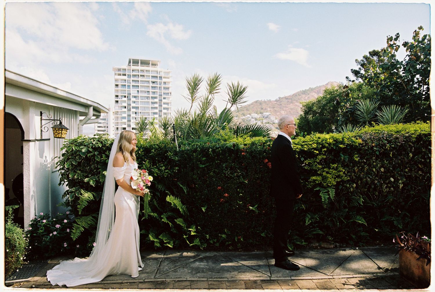Bride in white gown with veil holds bouquet during outdoor first look with groom at garden wedding venue.
