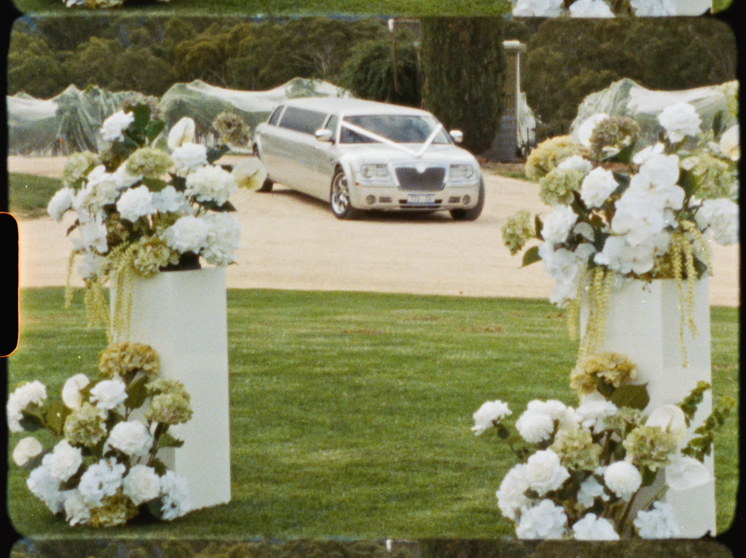 White floral pillars framing a silver stretch limousine arriving at an outdoor vineyard wedding.