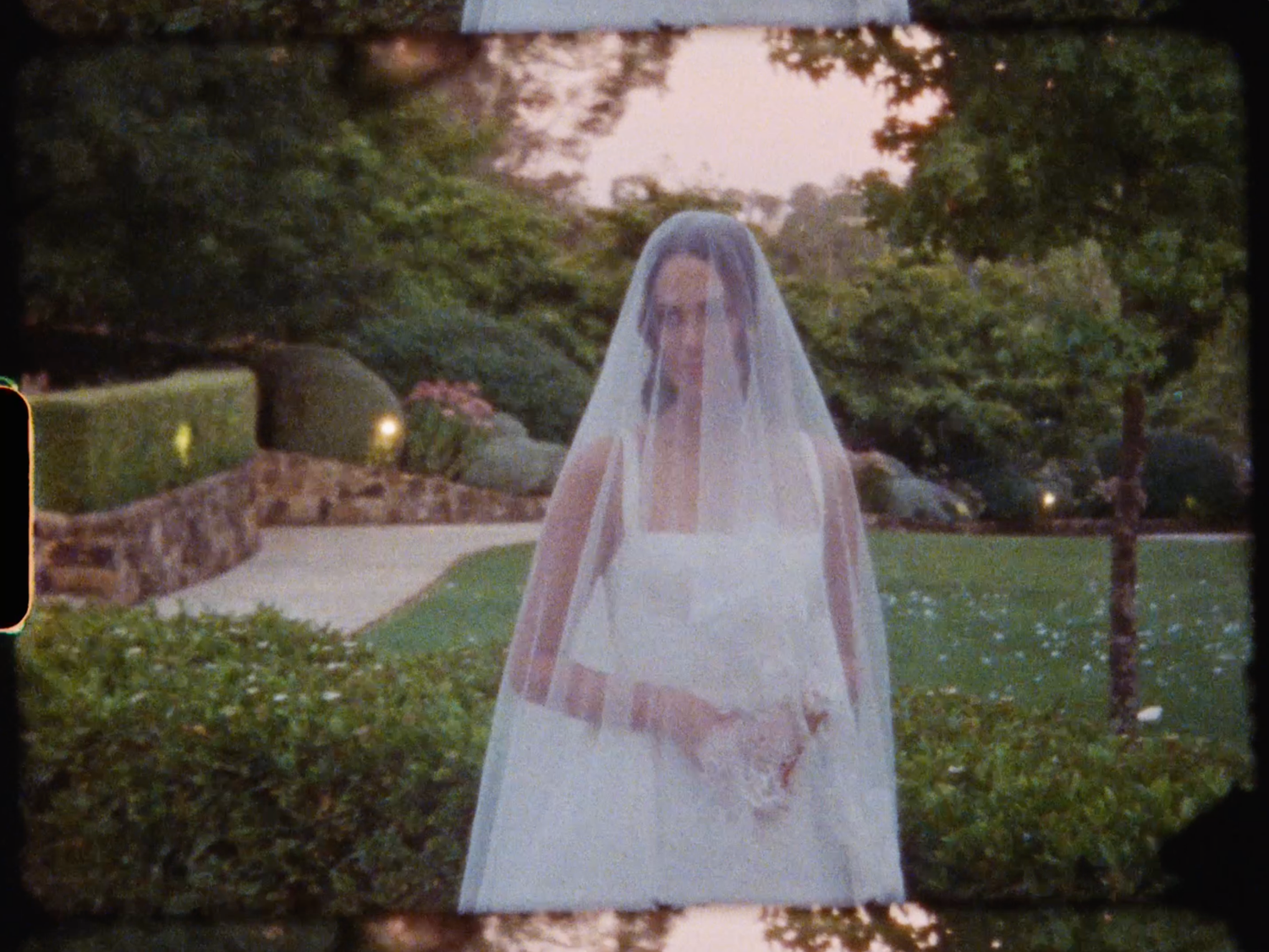 Bride in white gown and veil walks through a beautifully landscaped garden.