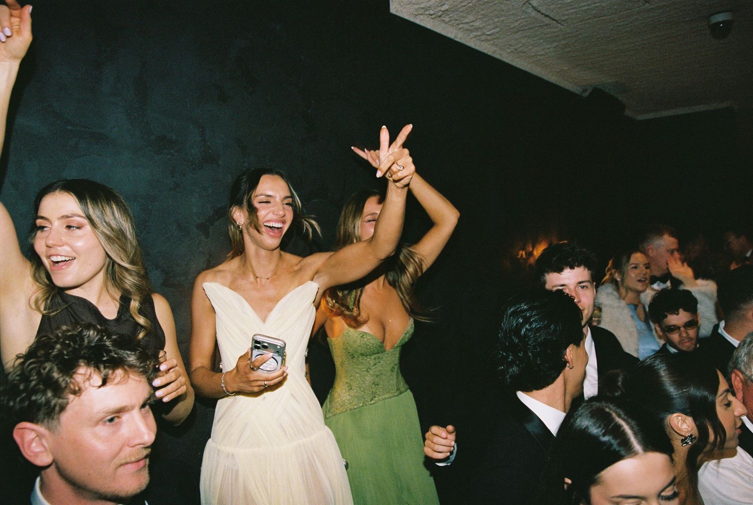 Bride in white dress dances joyfully with guests in green dress at lively wedding reception.