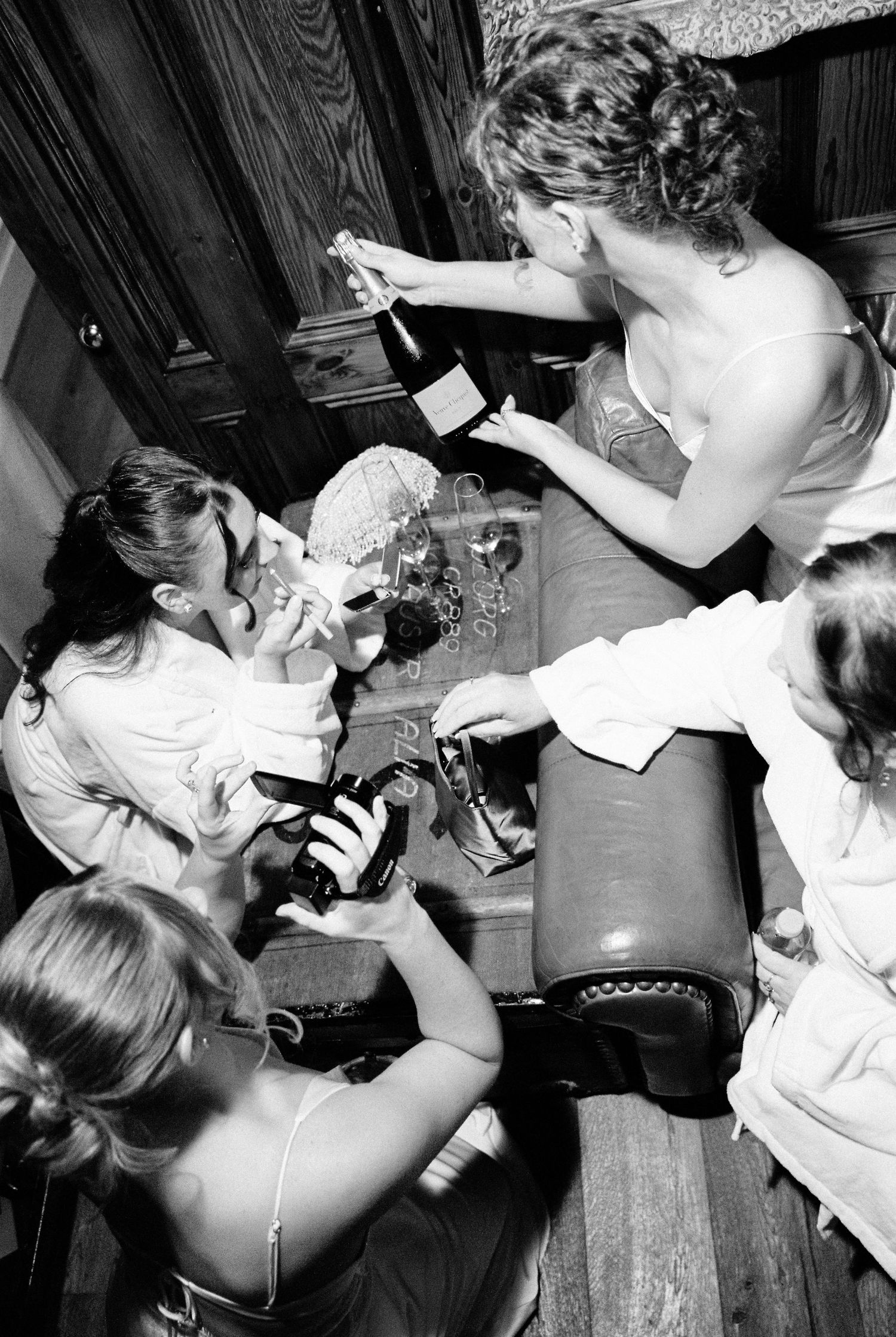 Bridesmaids gather around a table in black and white photo, sharing champagne during wedding preparations.