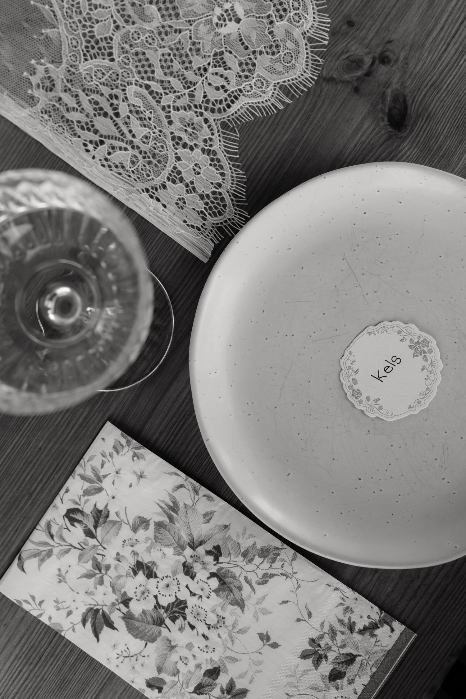 Black and white flat lay of wedding place setting with lace, ceramic plate, and floral menu card.