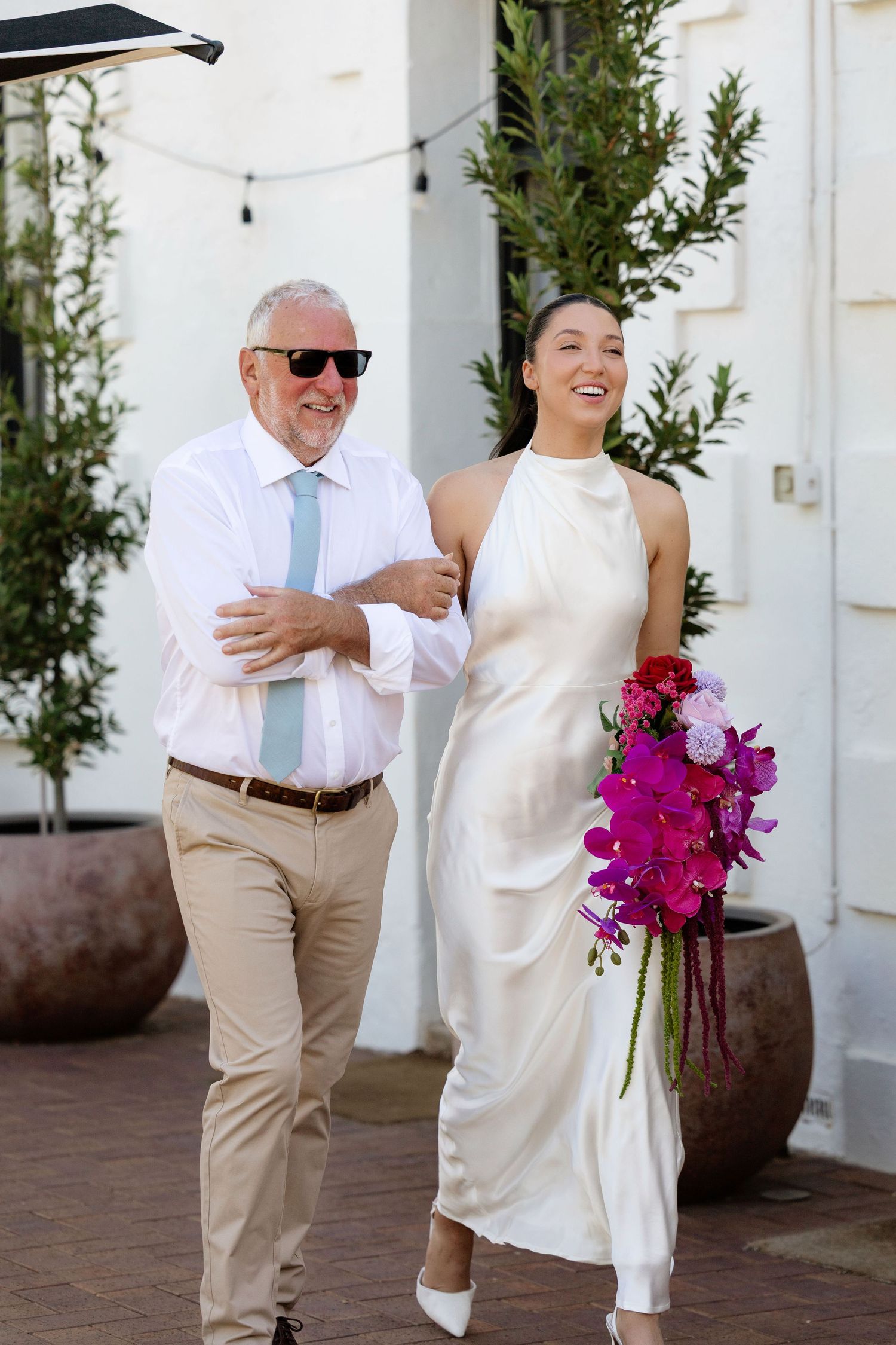 Bride in white halter gown walks arm-in-arm with older man in pink shirt, holding vibrant purple orchid bouquet.